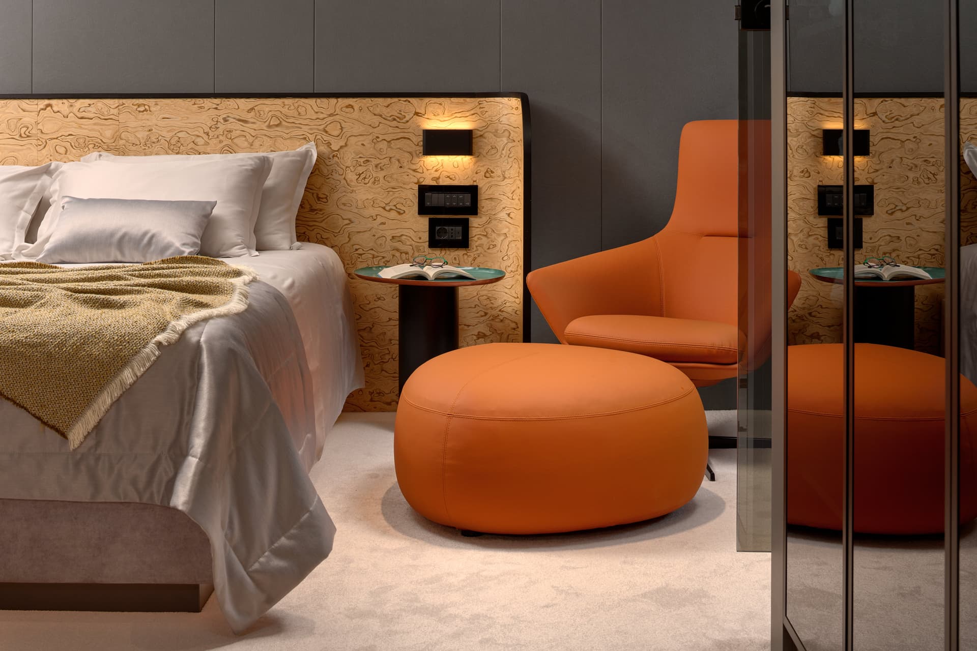 Bedroom interior with white bedding, orange armchair, ottoman, and wood-grain headboard panel.