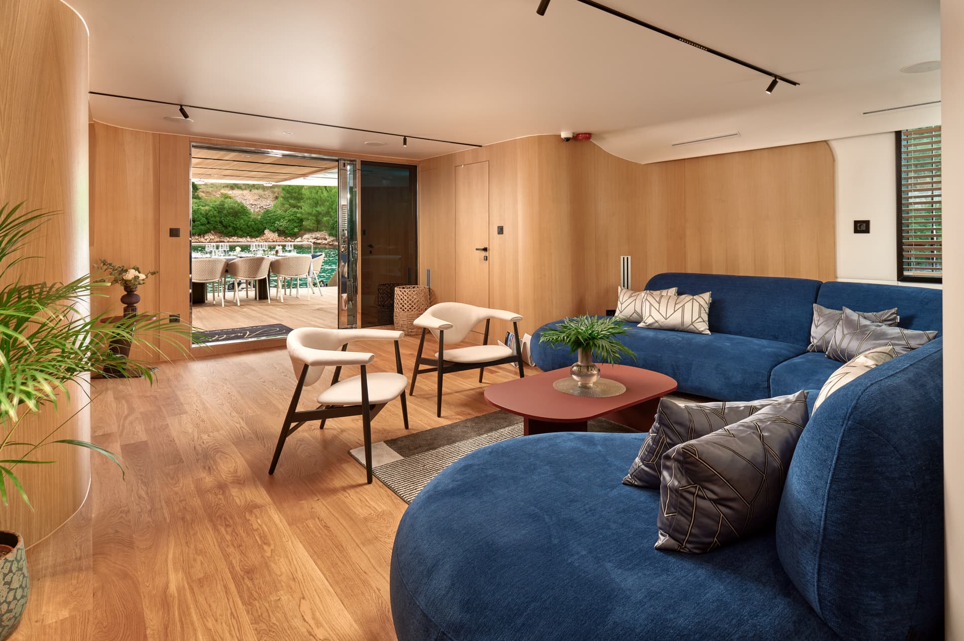 Modern yacht interior with blue sofa opening to an outdoor dining area near a rocky coast.