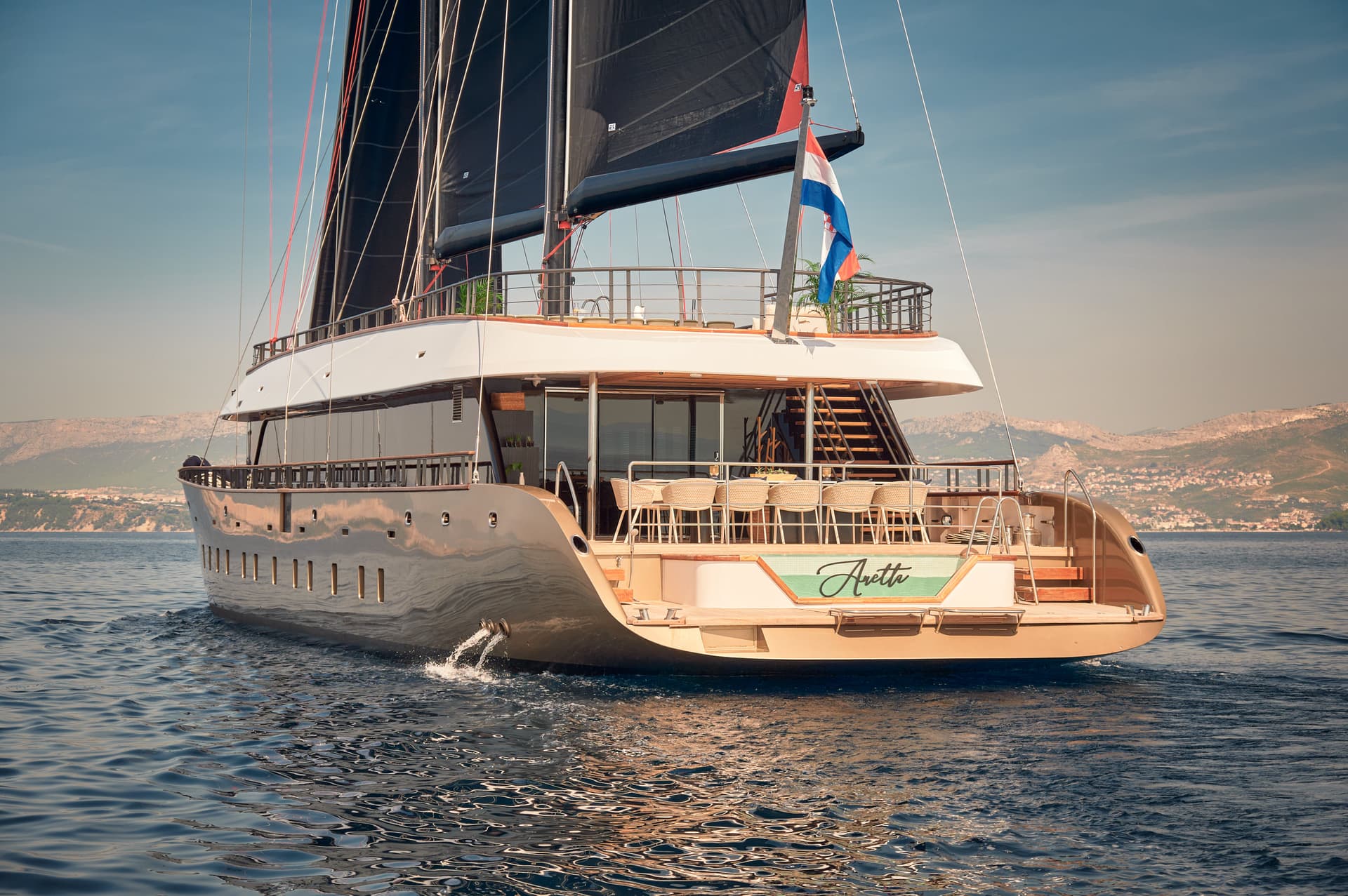 Luxury yacht Anetta sailing on blue water near a hilly coastline with a coastal town.