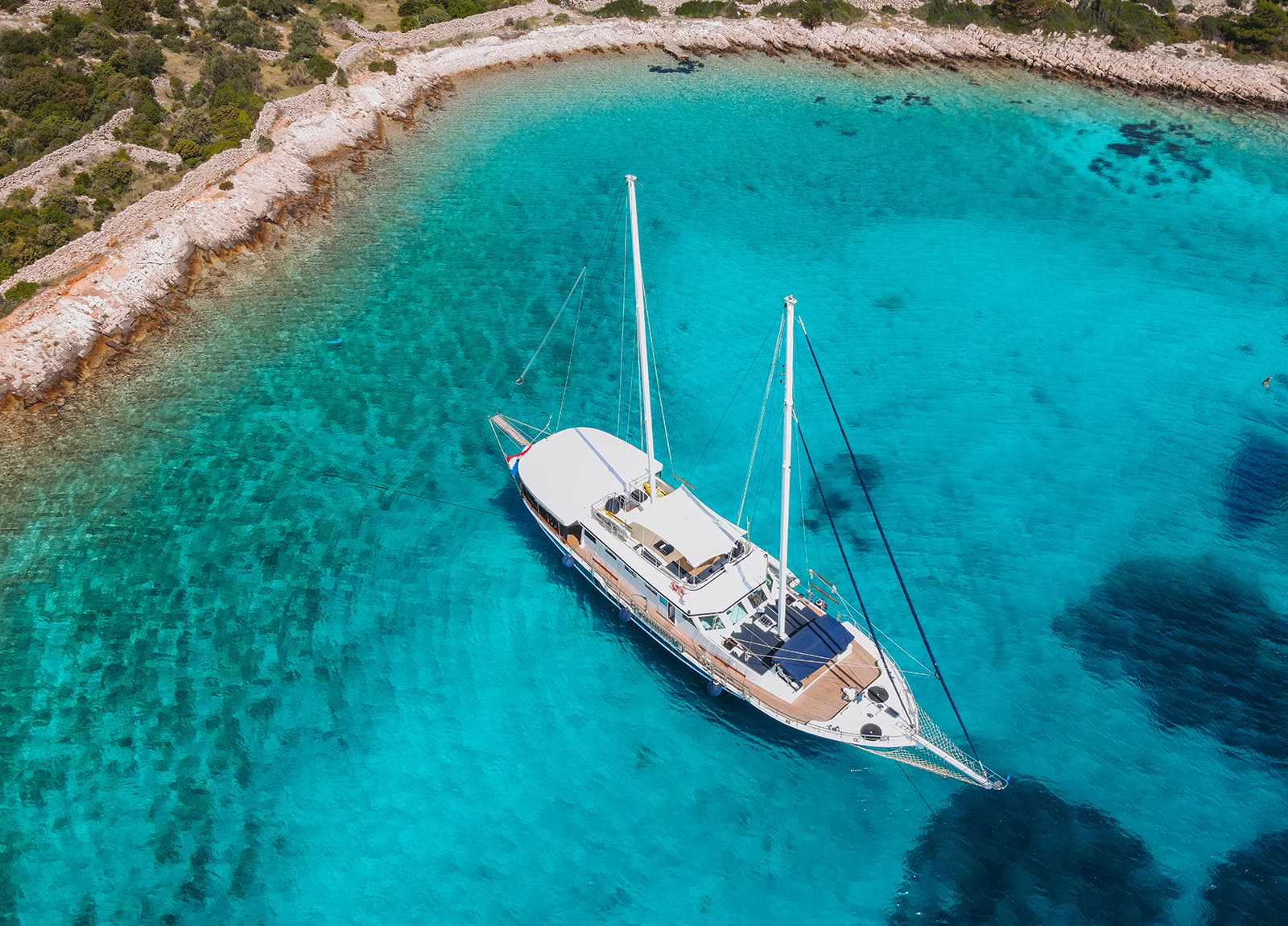 Large sailboat anchored in clear turquoise cove near rocky, scrub-covered coastline.