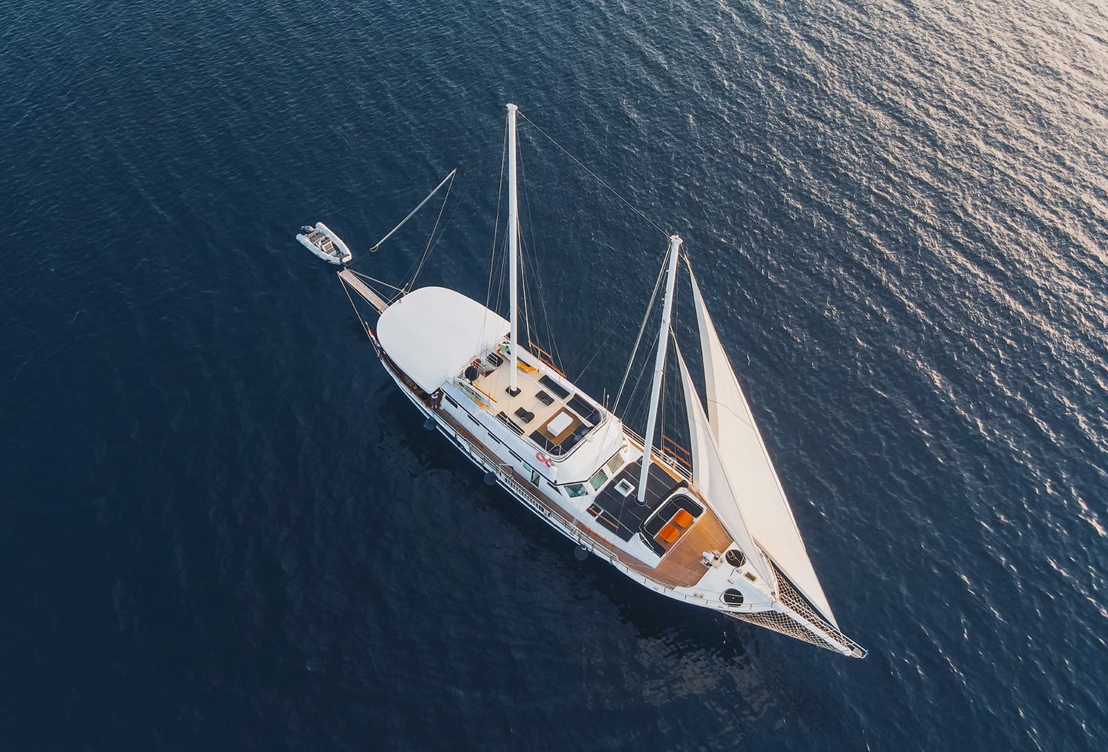 Large white sailboat with sails up and a dinghy alongside on deep blue water.