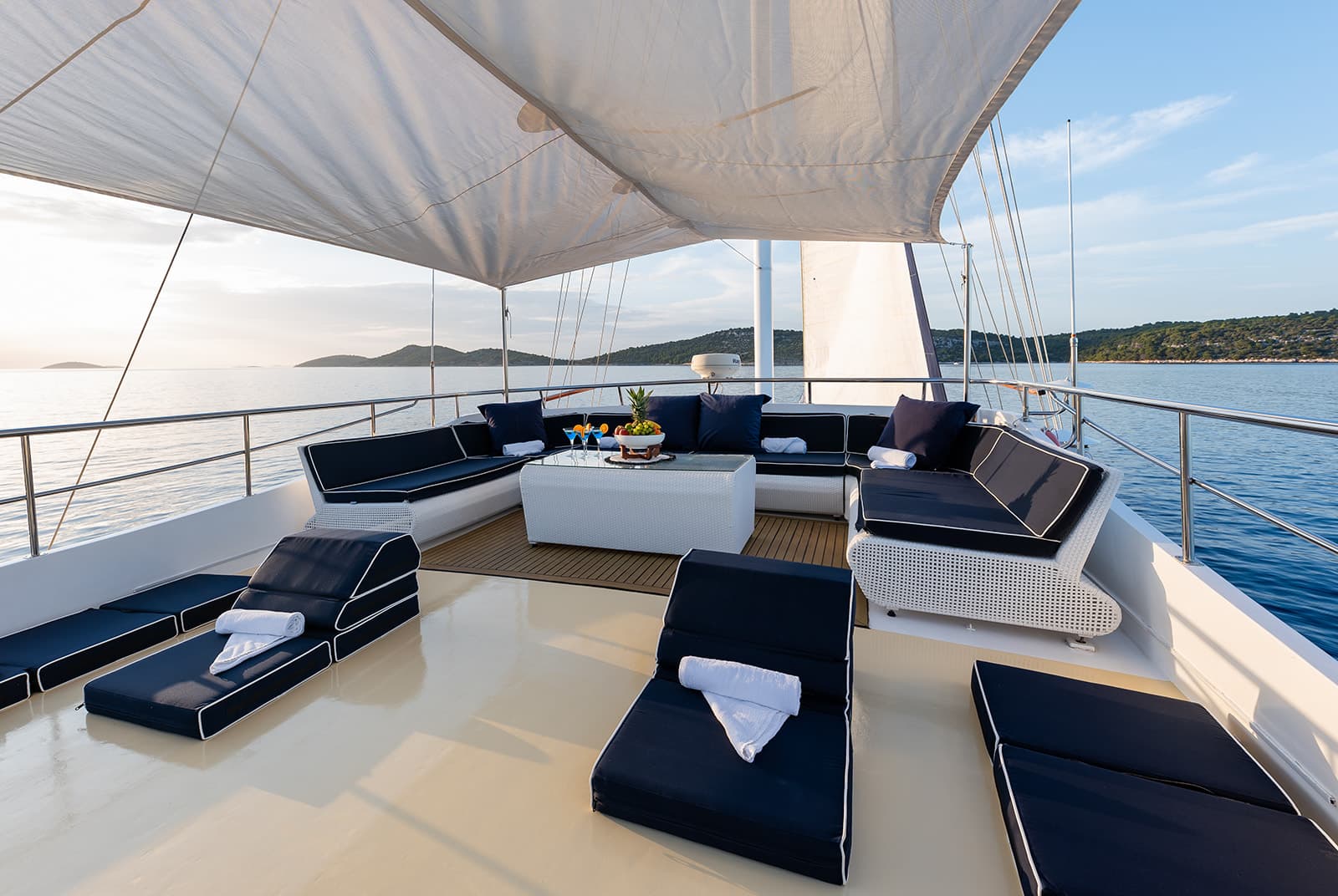 Yacht deck with white awning, navy cushions, and fruit platter cruising near hilly coastline.