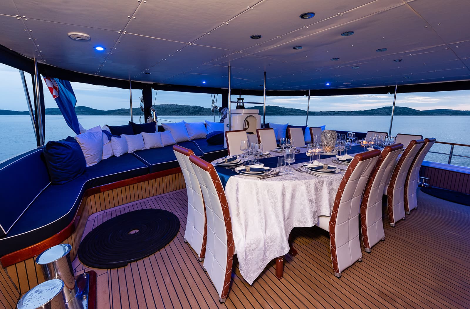 Formal dining setup on yacht deck with blue cushions, overlooking islands at dusk.