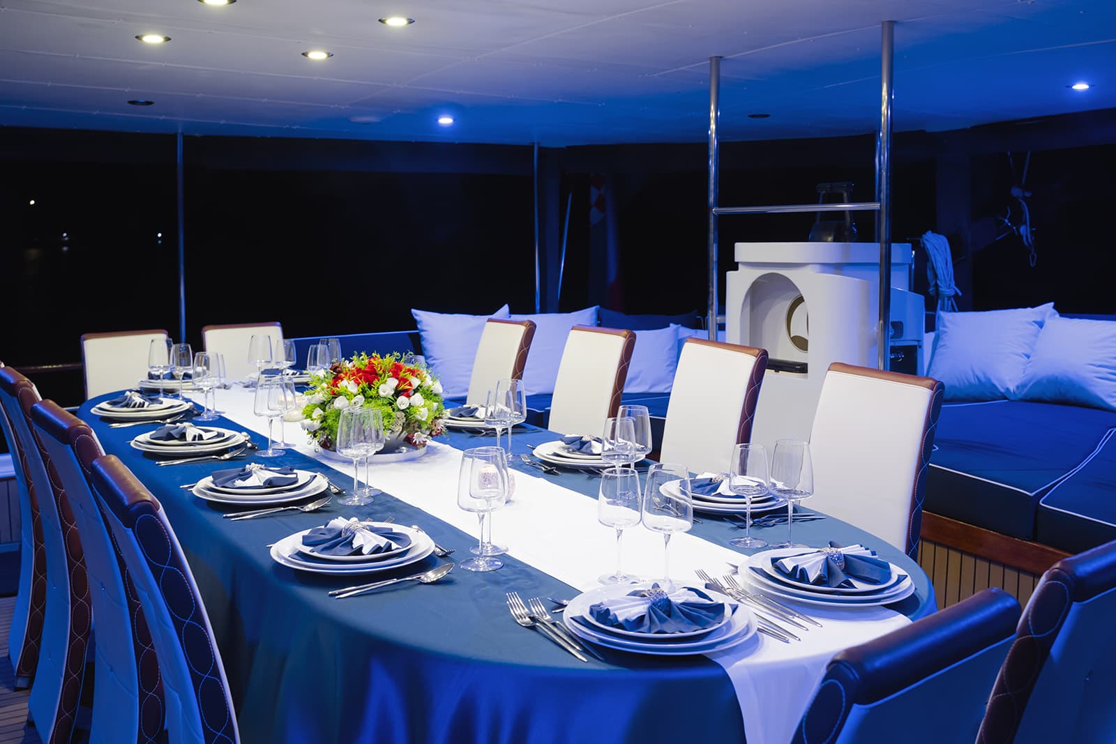 Formal dining table set for many guests on a yacht deck at night with blue lighting.