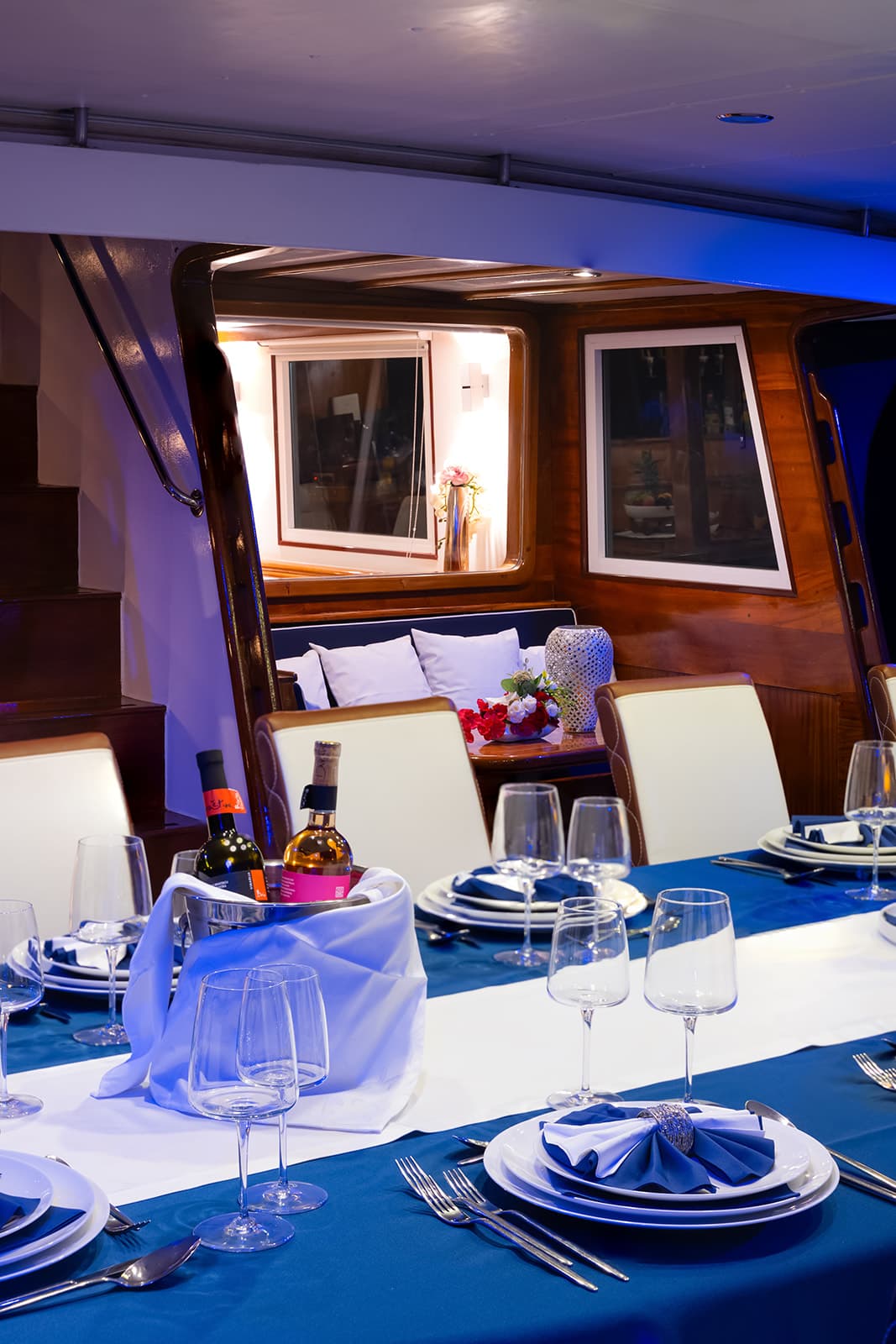 Formal dining table set inside a yacht cabin with blue and white decor and wine bottles.