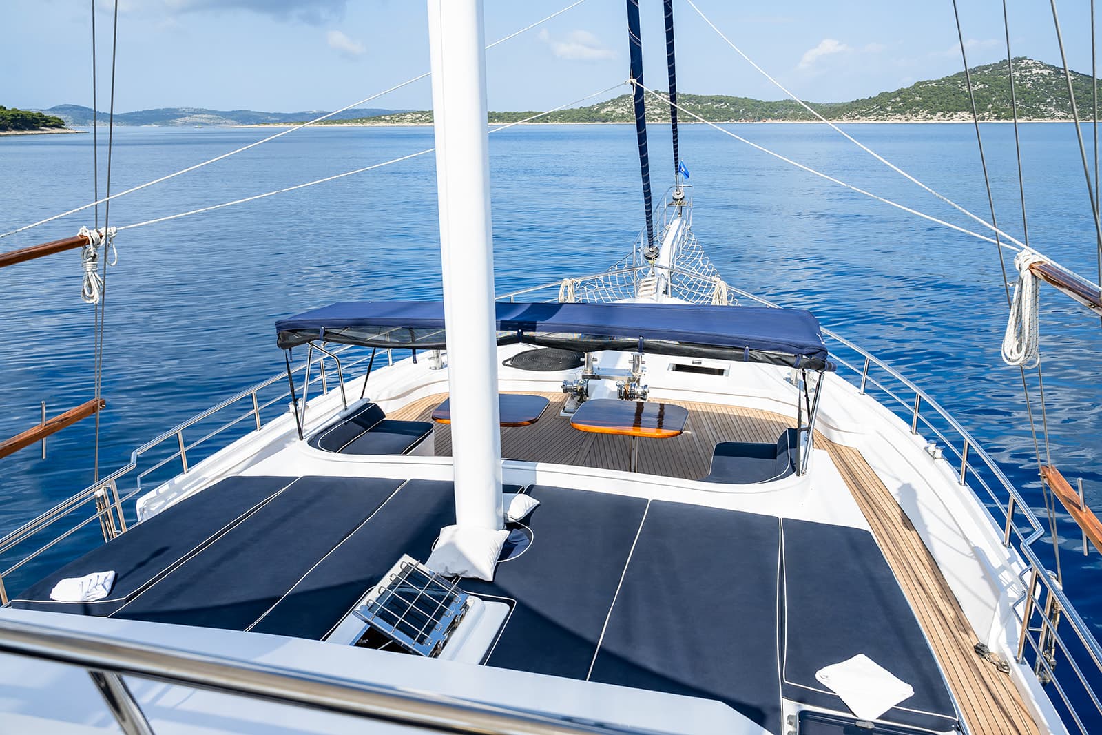View from bow of sailboat on blue sea toward hilly, green coastline.