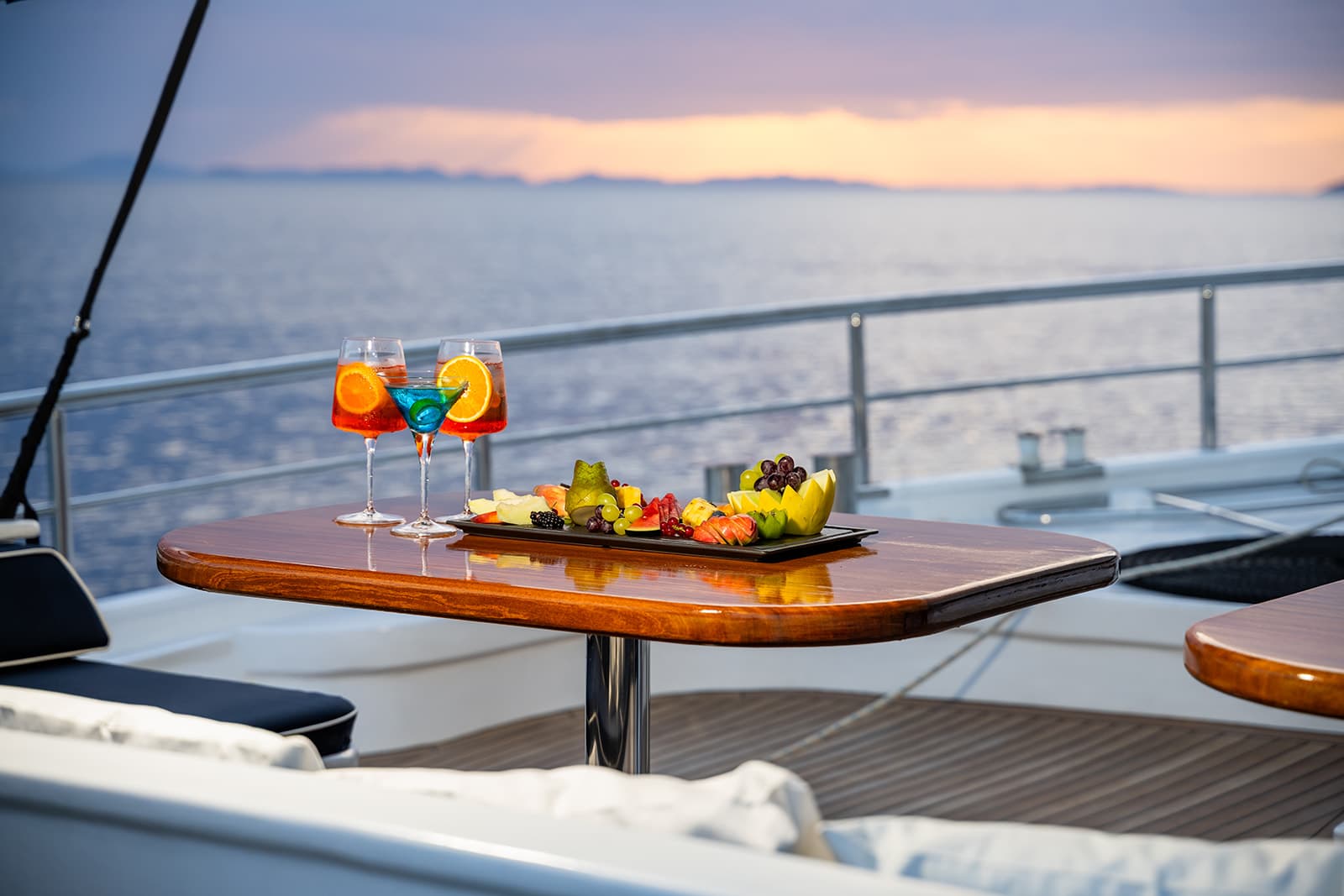 Cocktails and fresh fruit platter on a glossy table aboard a yacht at sunset over the sea.