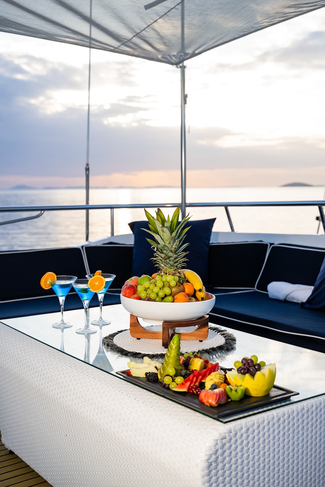 Fruit platter and blue cocktails on a glass table aboard a boat at sunset