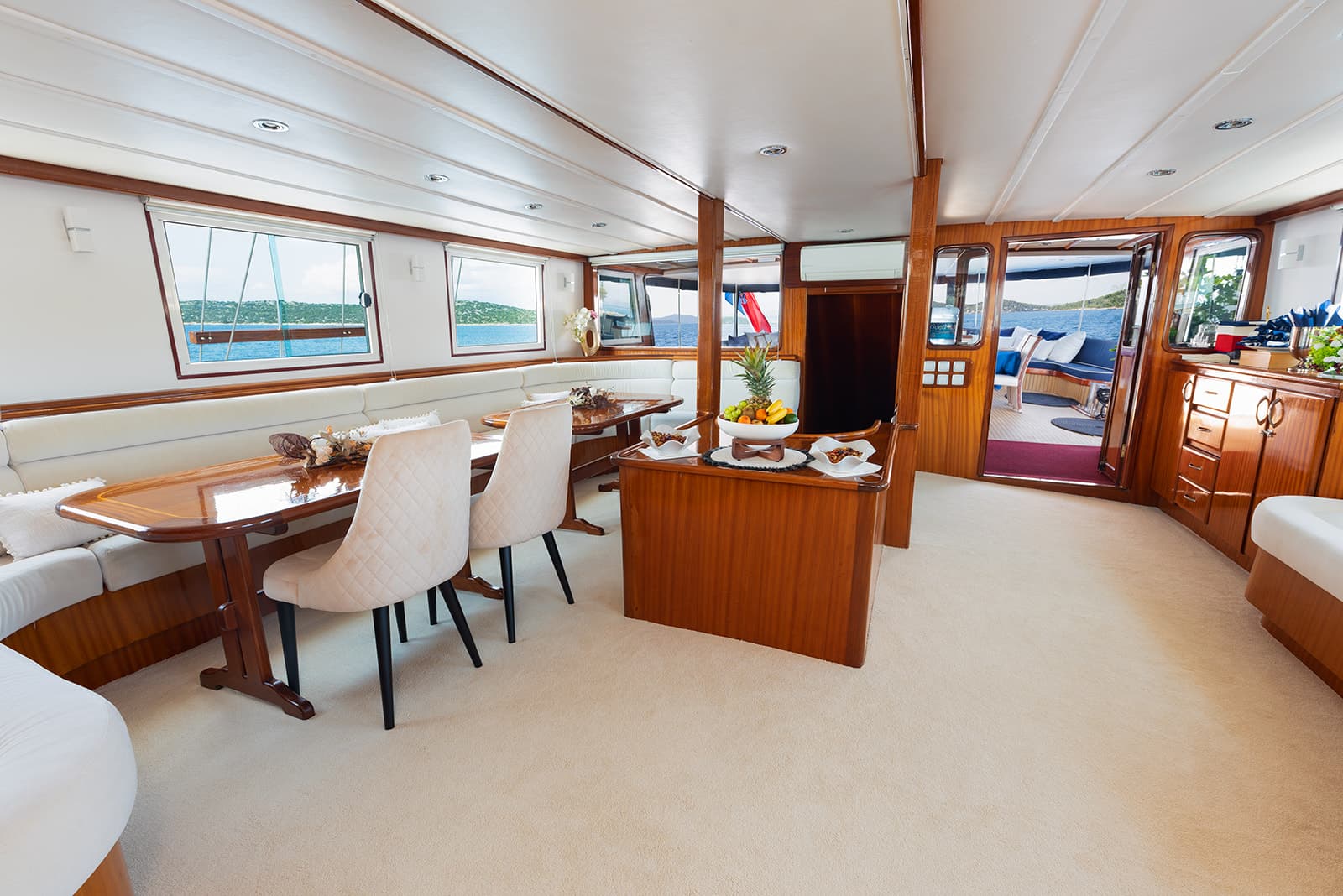 Yacht interior dining area with wood trim, white seating, and view of blue water and green islands.