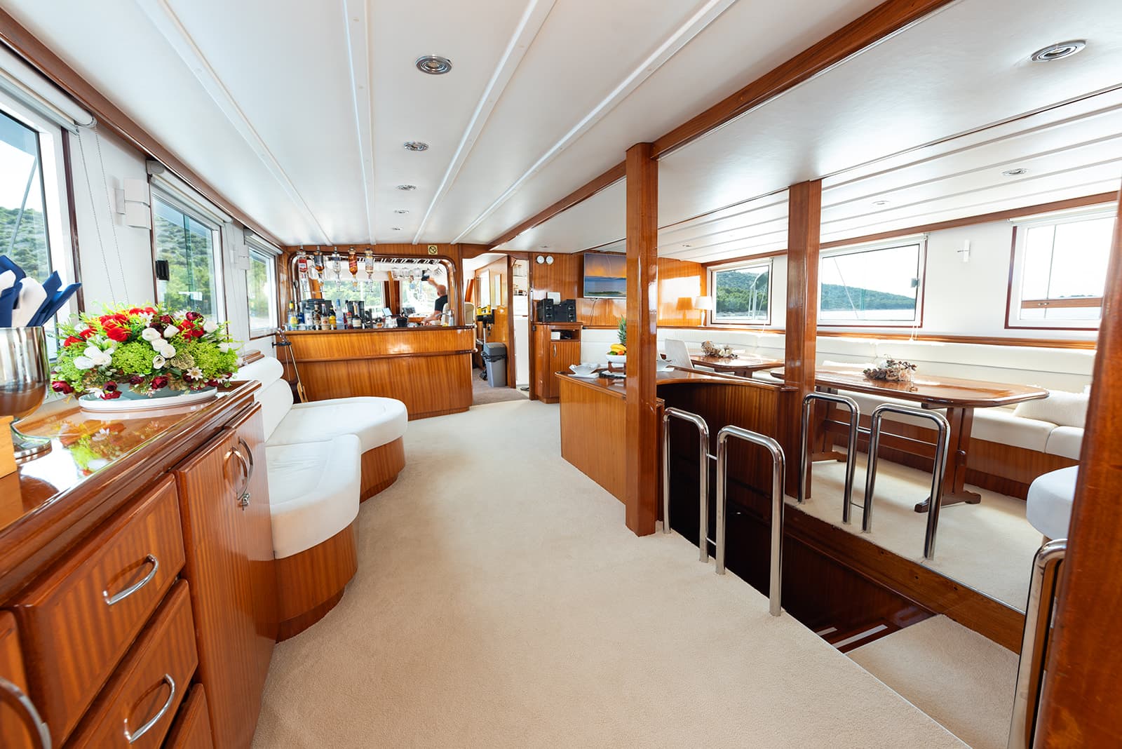 Interior salon of a yacht with wood paneling, white seating, a bar, and views of green hillsides.