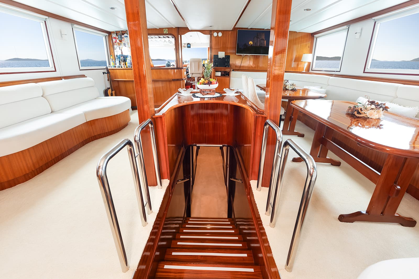 Yacht interior with polished wood, white seating, and stairs leading below deck, sea visible.