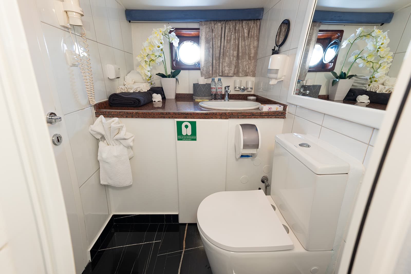 Small white bathroom with toilet, sink, mirror, and porthole window suggesting a boat cabin.