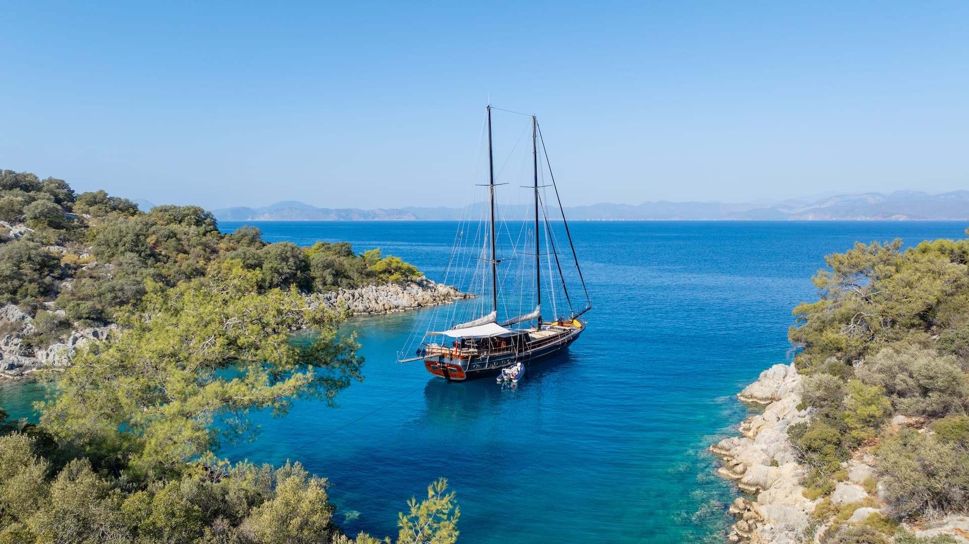 Two-masted wooden gulet anchored in bright blue water of a hidden bay with rocky, green coastline.