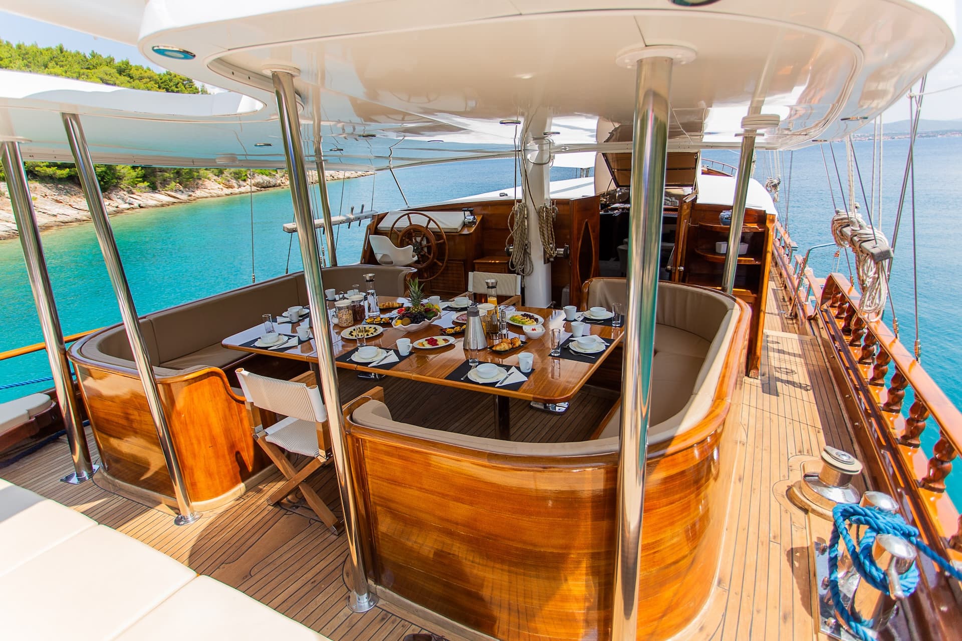 Dining table set for breakfast on a wooden sundeck of a boat near a forested coastline.