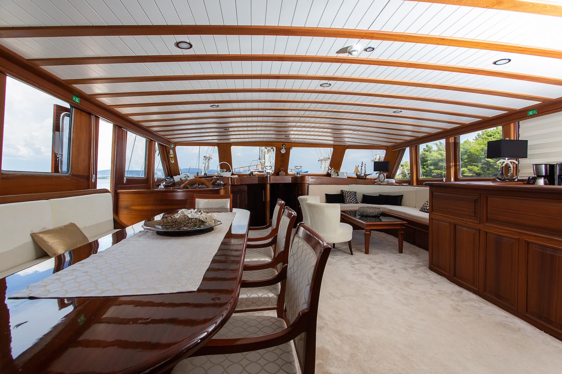Yacht interior dining area with wood trim, white ceiling, and views of trees and water.