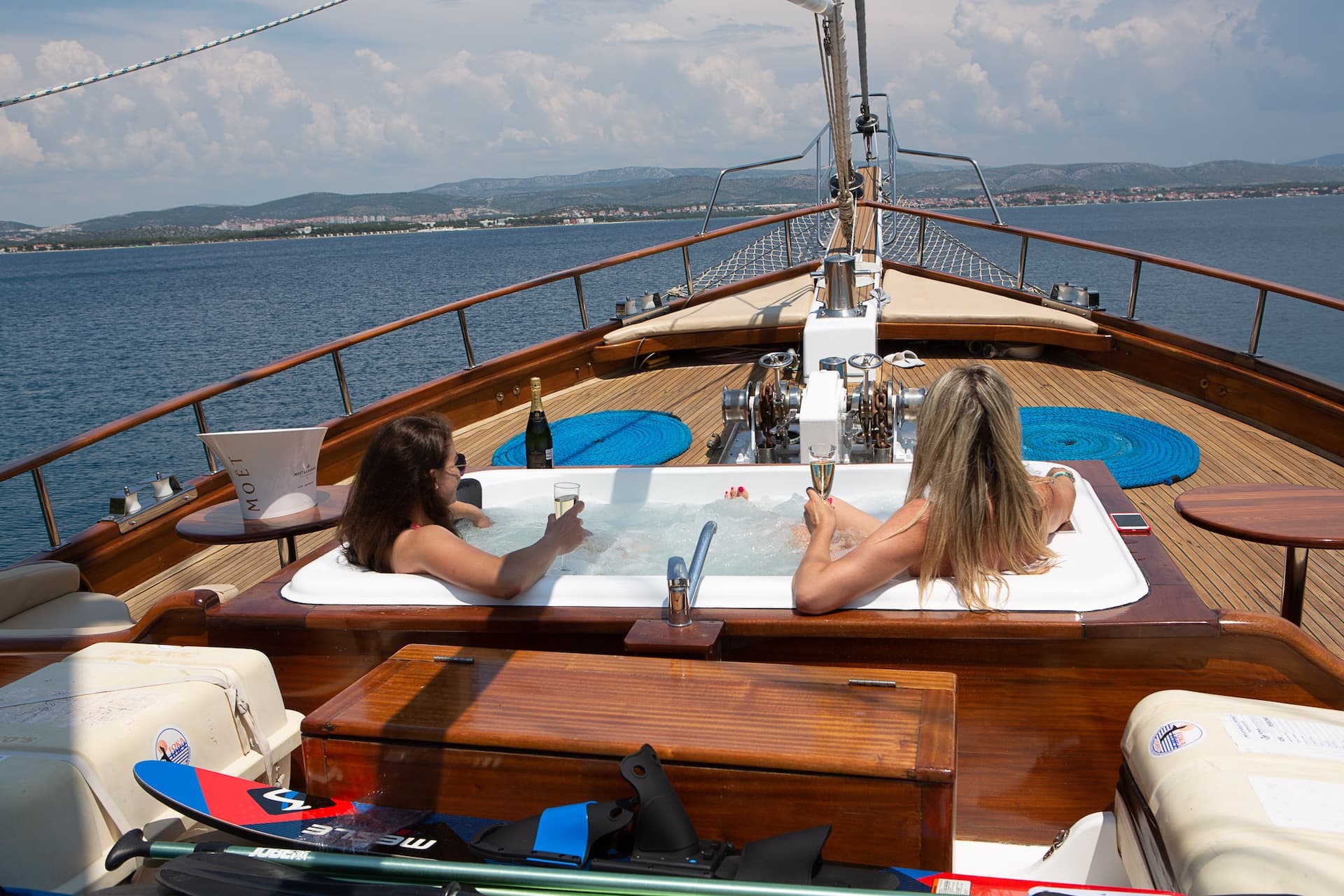 Women relaxing in a boat jacuzzi with champagne overlooking a coastal town and hills.