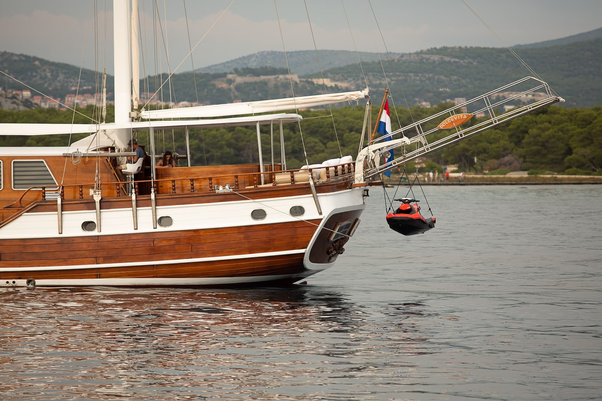 Wooden boat named Dolce Vita with jet ski suspended off the bow near a forested coastline.