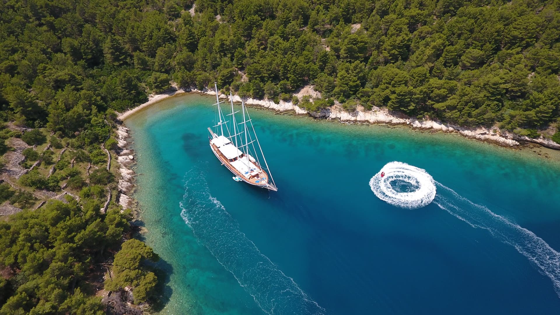 Sailboat anchored near forested coastline while someone tows a circular wake on turquoise sea.