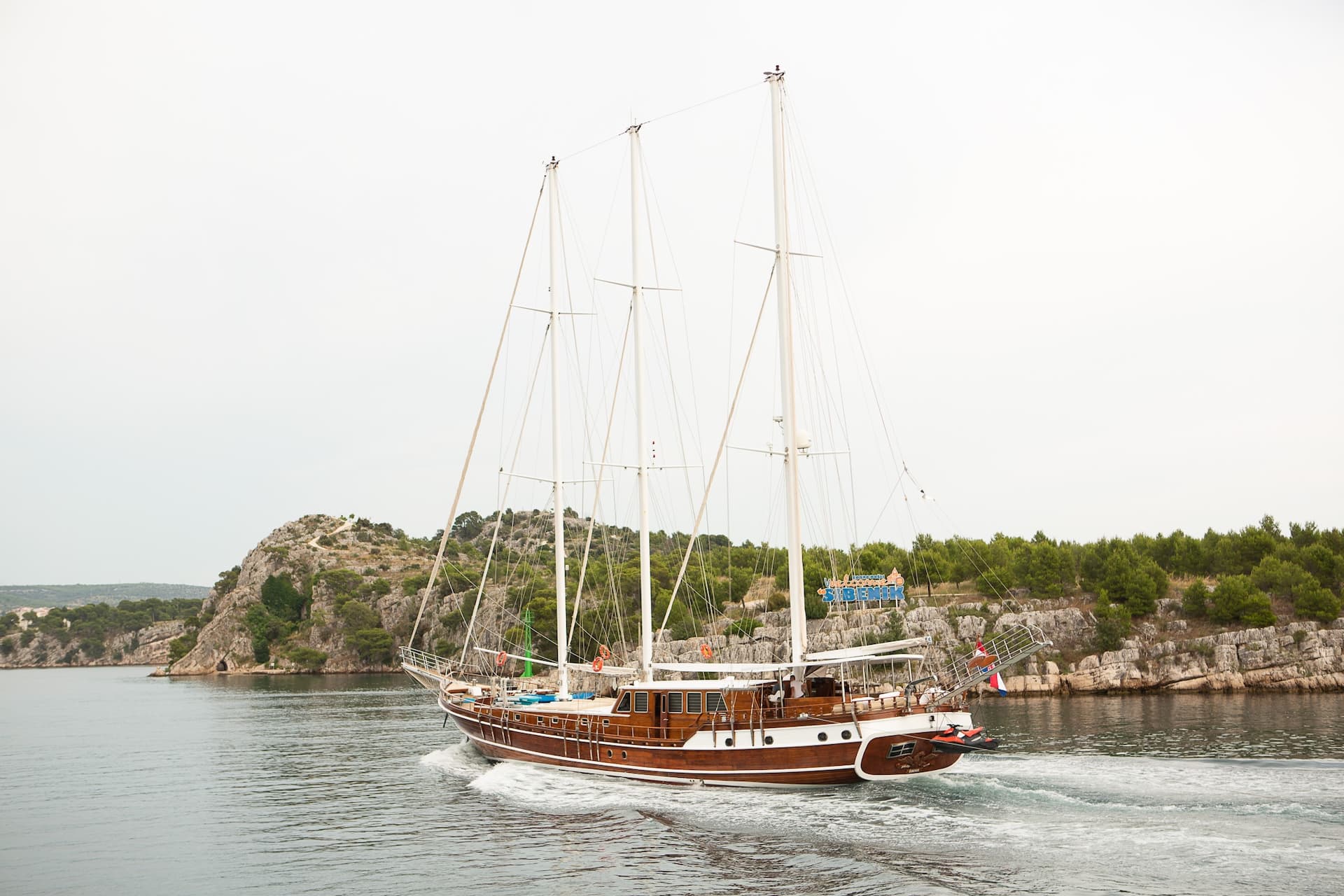 Wooden gulet sailing past rocky, tree-covered coastline on cloudy day