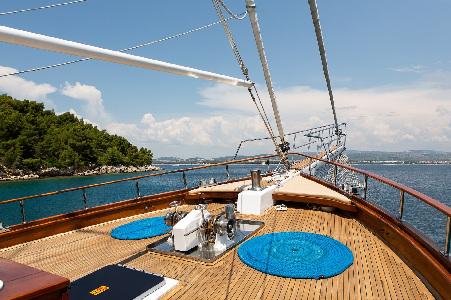 Bow of wooden yacht with winch and blue rope sailing near forested coastline under blue sky.