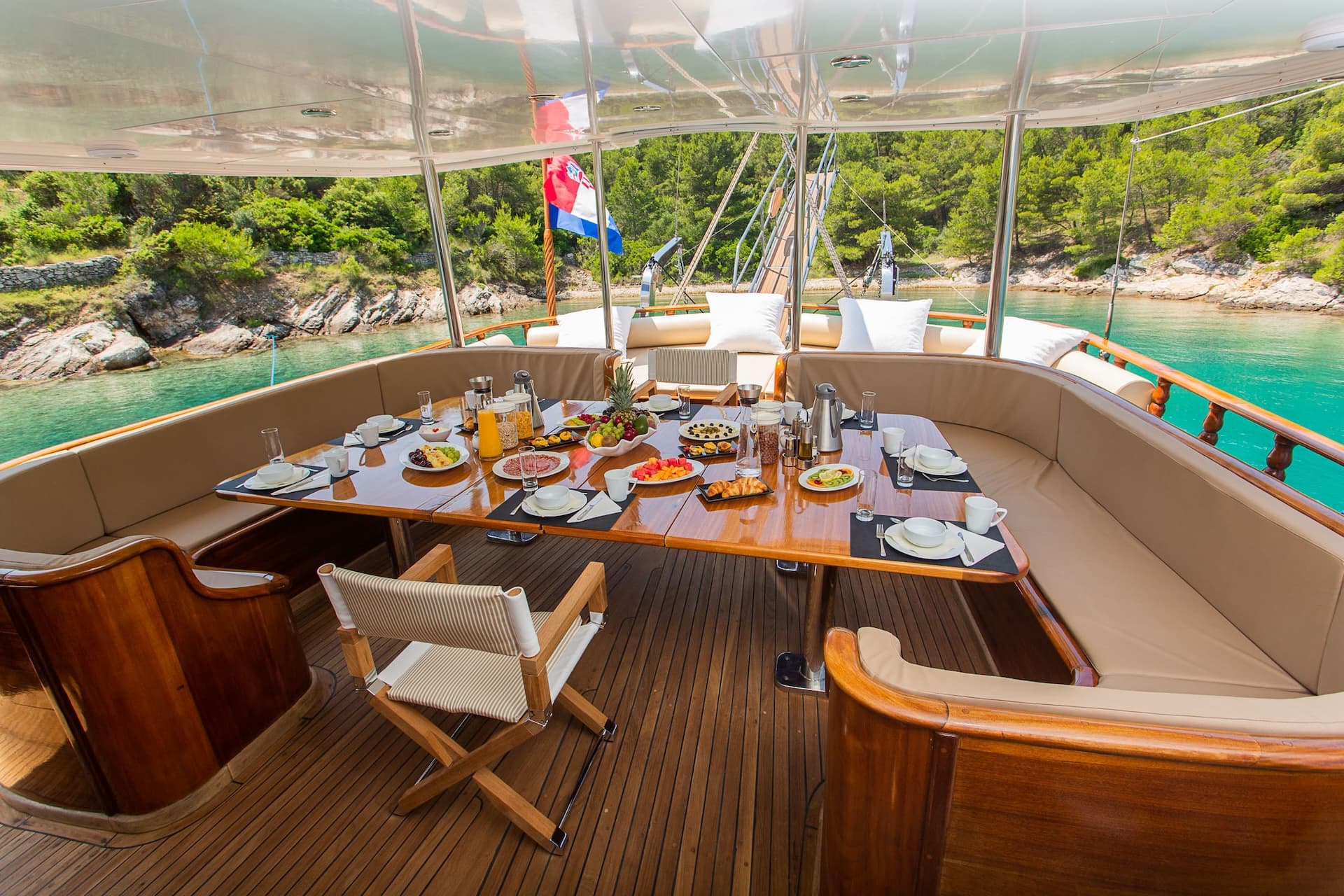 Breakfast table set on a yacht sundeck anchored near a lush, rocky coastline.