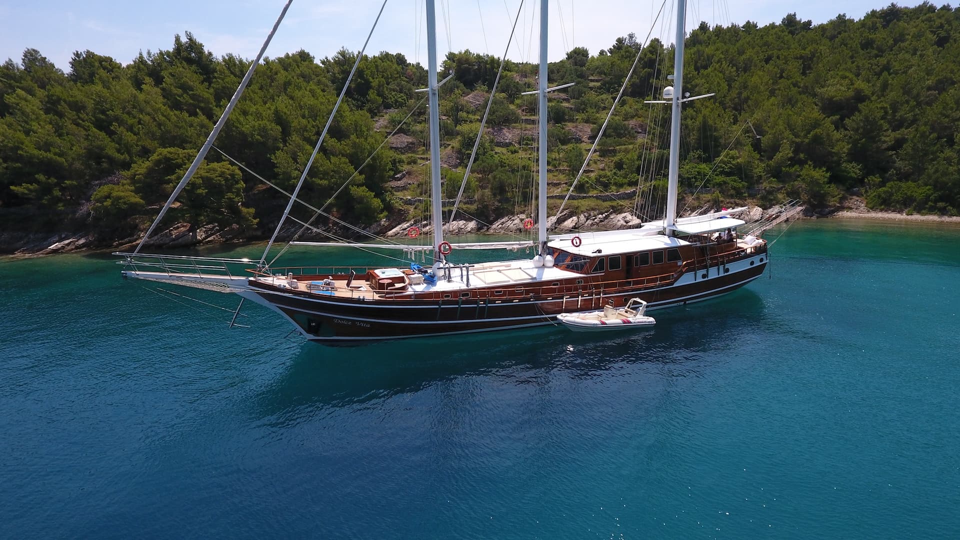 Wooden sailboat named Dolce Vita anchored in clear turquoise water near forested coastline.
