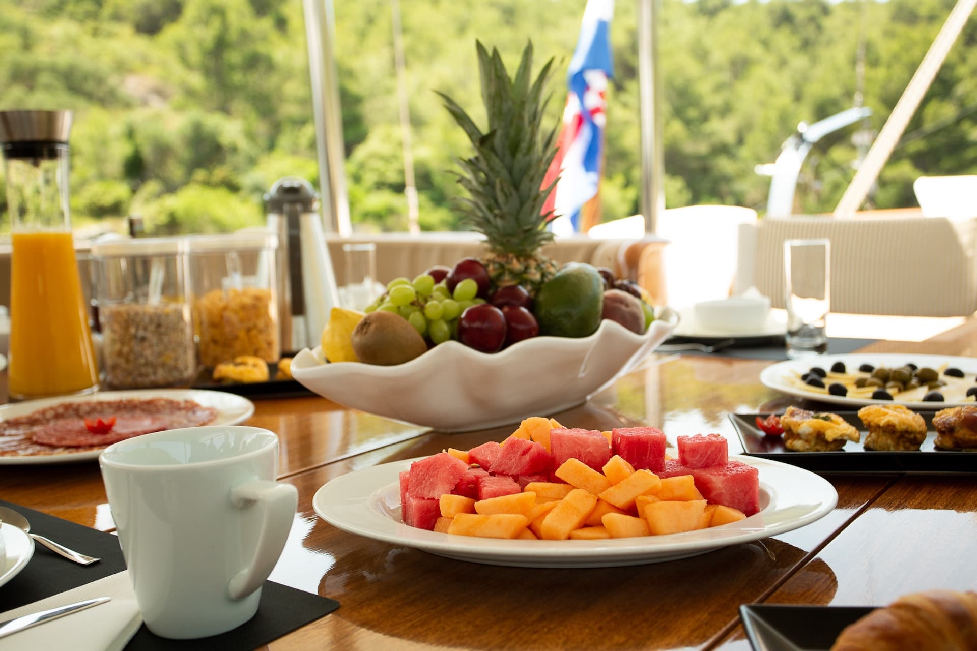 Breakfast spread with fruit, juice, and cold cuts on a table aboard a vessel with lush greenery outside.