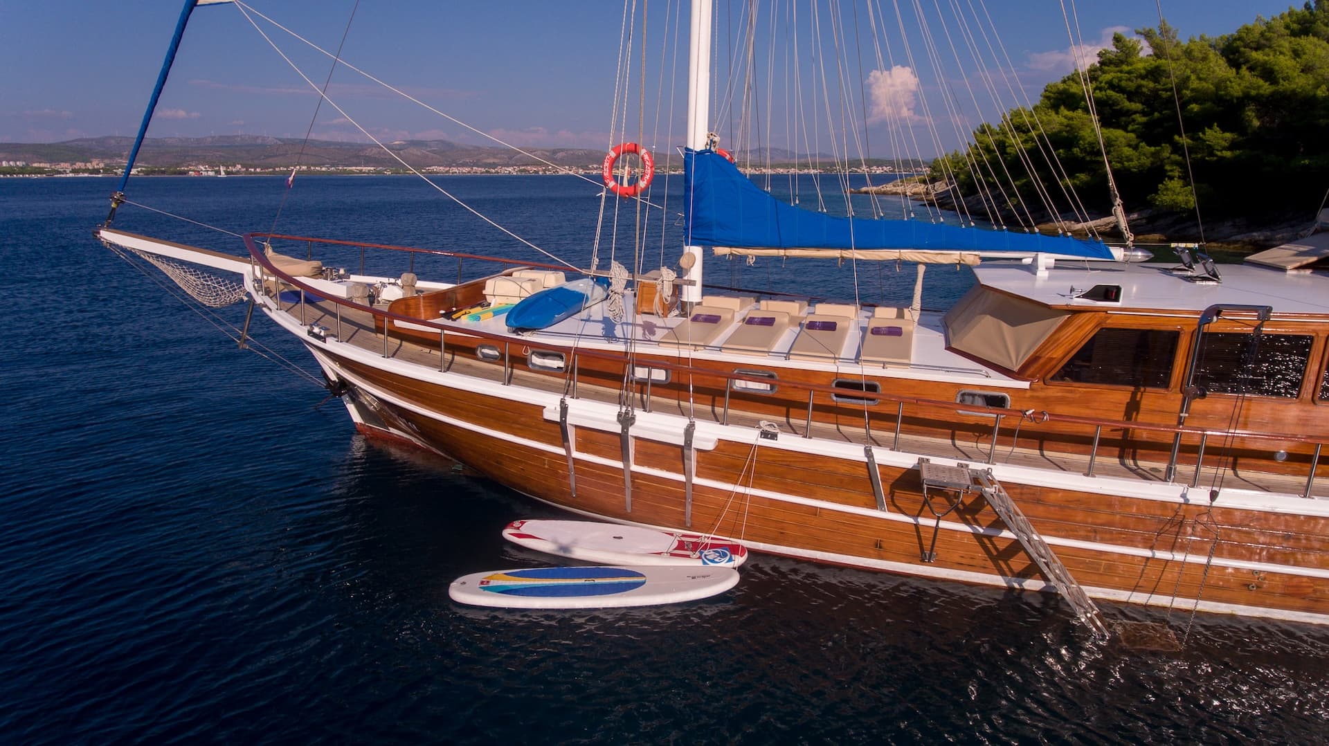 Wooden gulet Malena boat moored near a wooded shore with paddleboards in dark blue water.