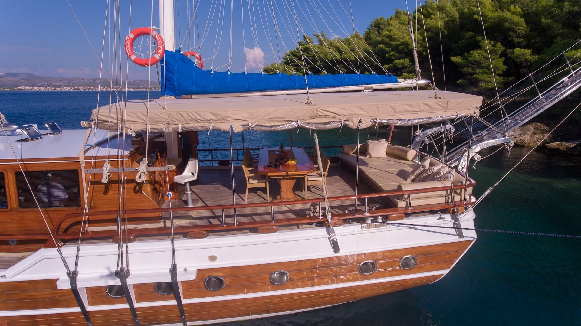 Wooden gulet boat deck with dining table and sunbeds anchored near green coastline.