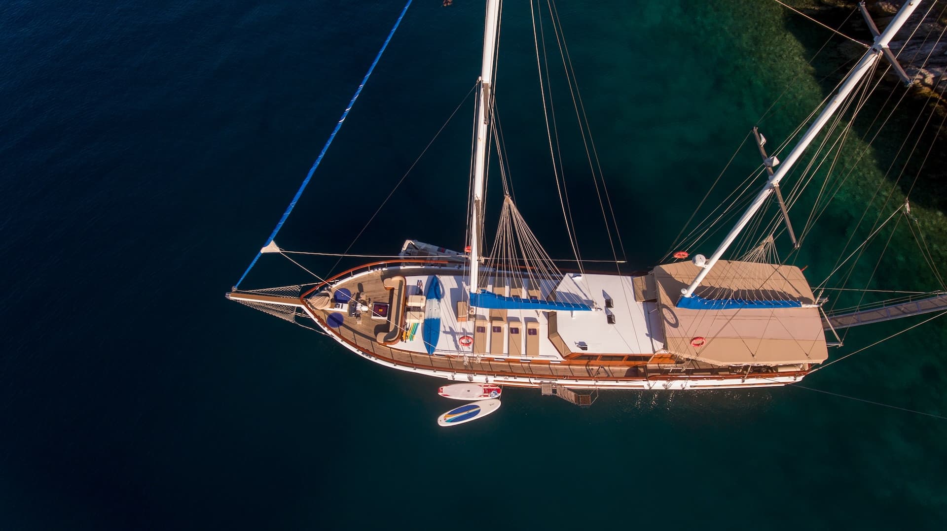 Aerial view of a large sailboat anchored in clear, dark blue coastal water near rocks.
