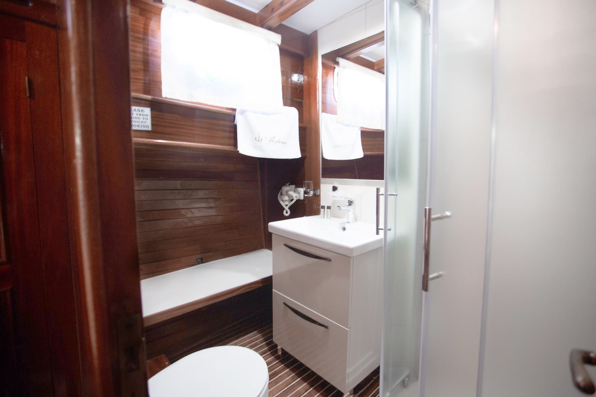 Compact bathroom with dark wood paneling, white vanity, and frosted glass shower stall on a boat.