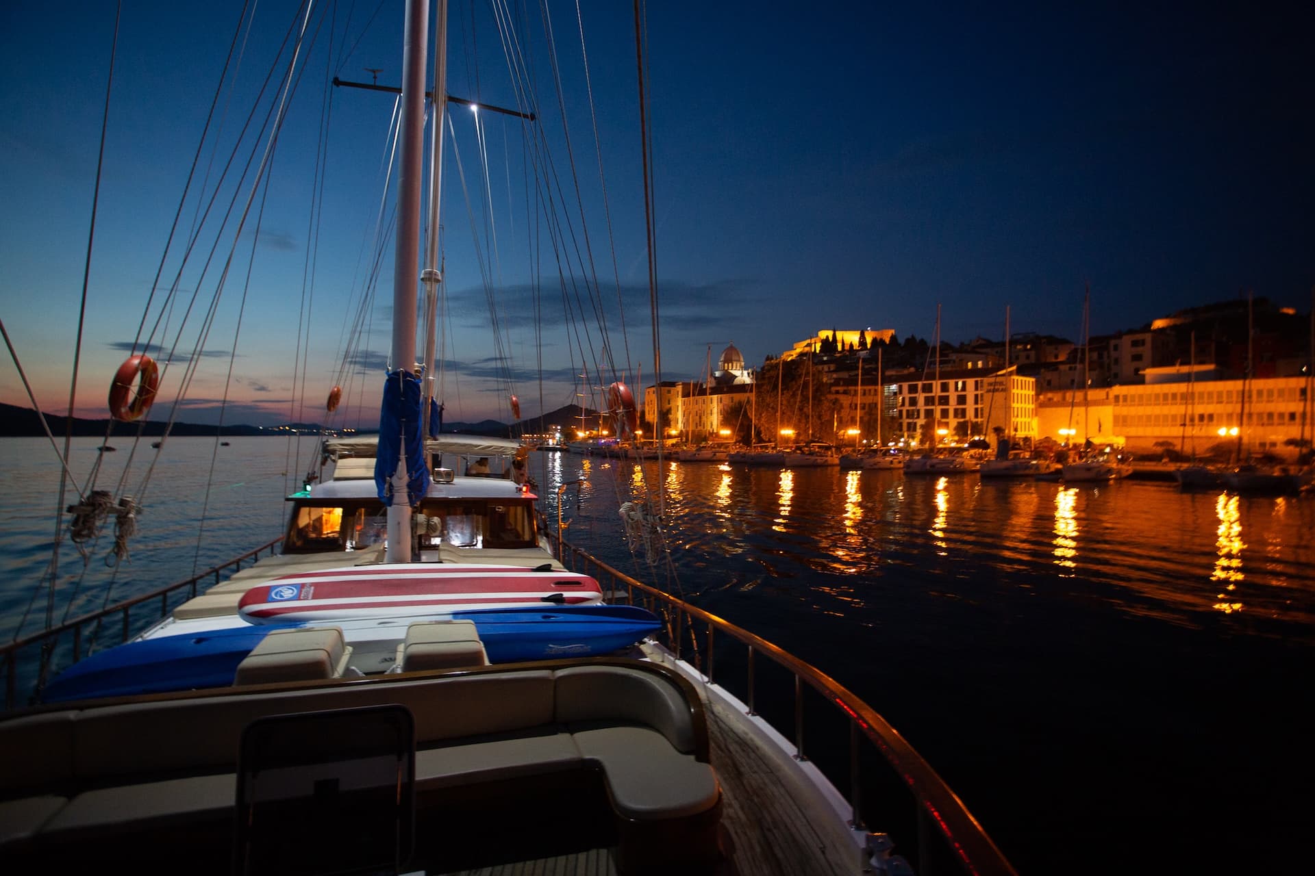 View from sailboat deck toward illuminated harbor town at twilight with docked boats.
