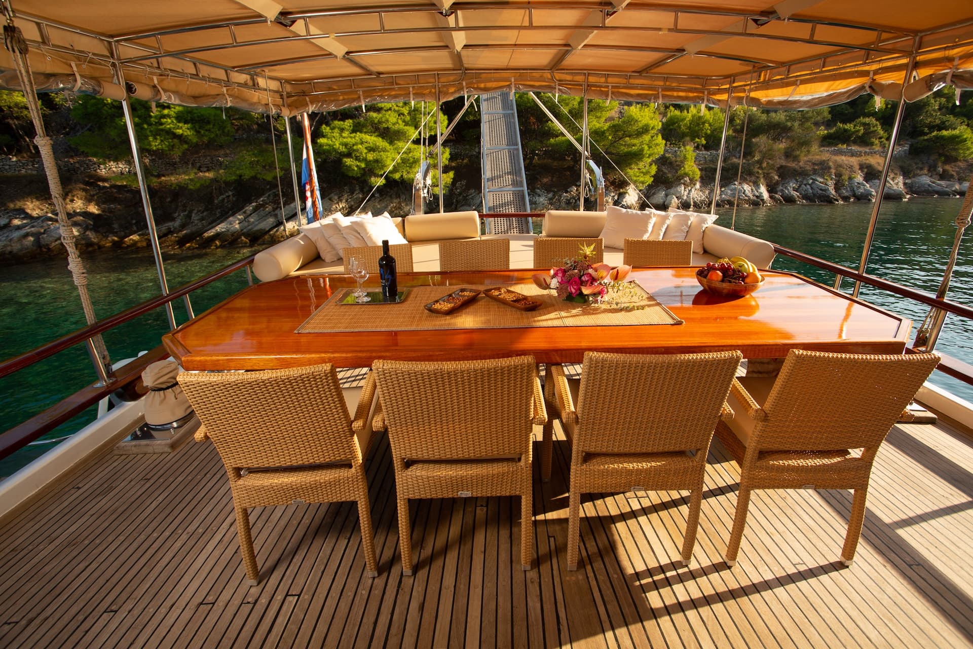 Dining table set on teak deck of boat anchored near rocky, forested coastline.