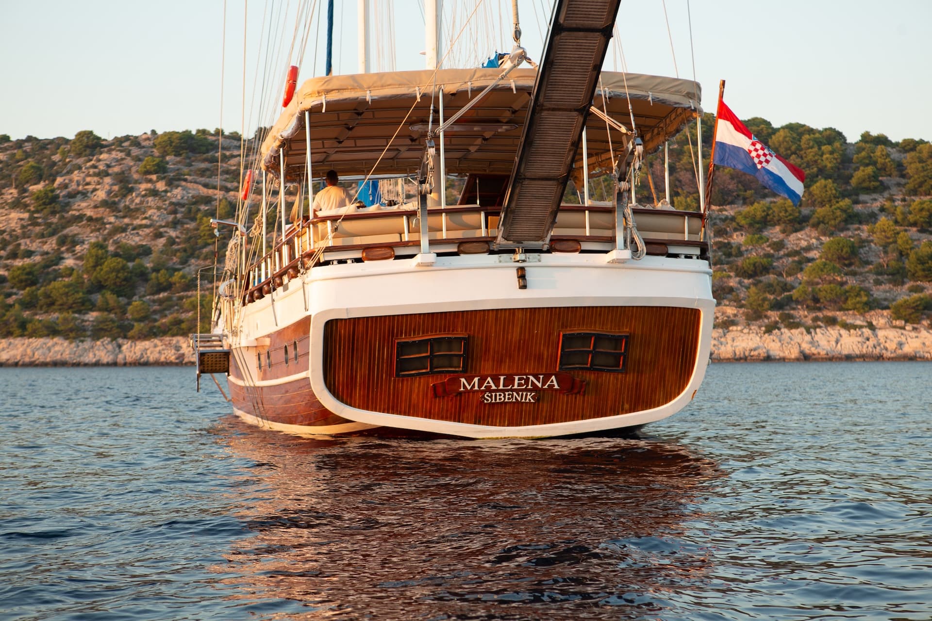 Malena boat from Šibenik with Croatian flag sailing near rocky, tree-covered coastline.