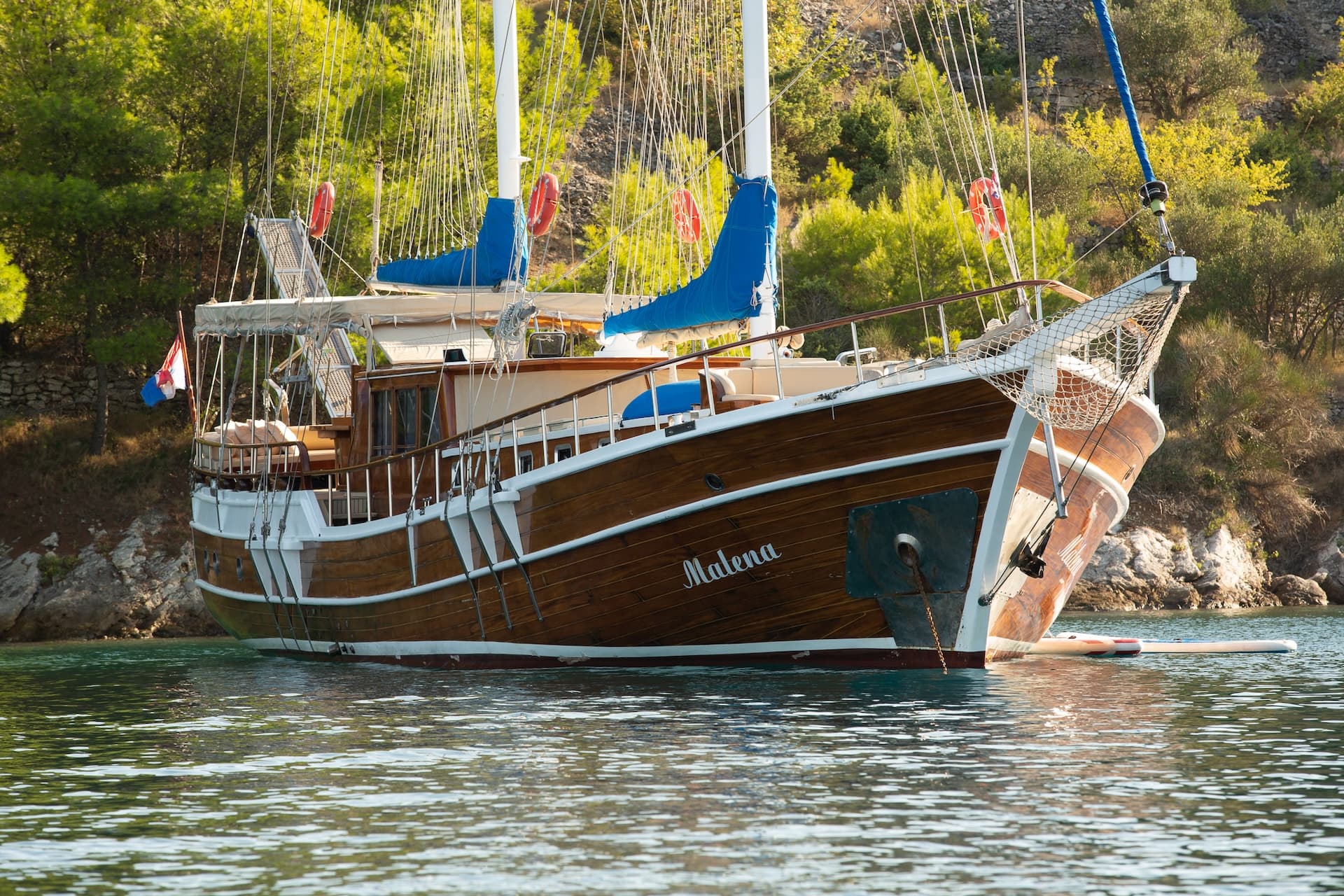 Wooden sailboat named Malena anchored near a rocky, tree-covered coastline.