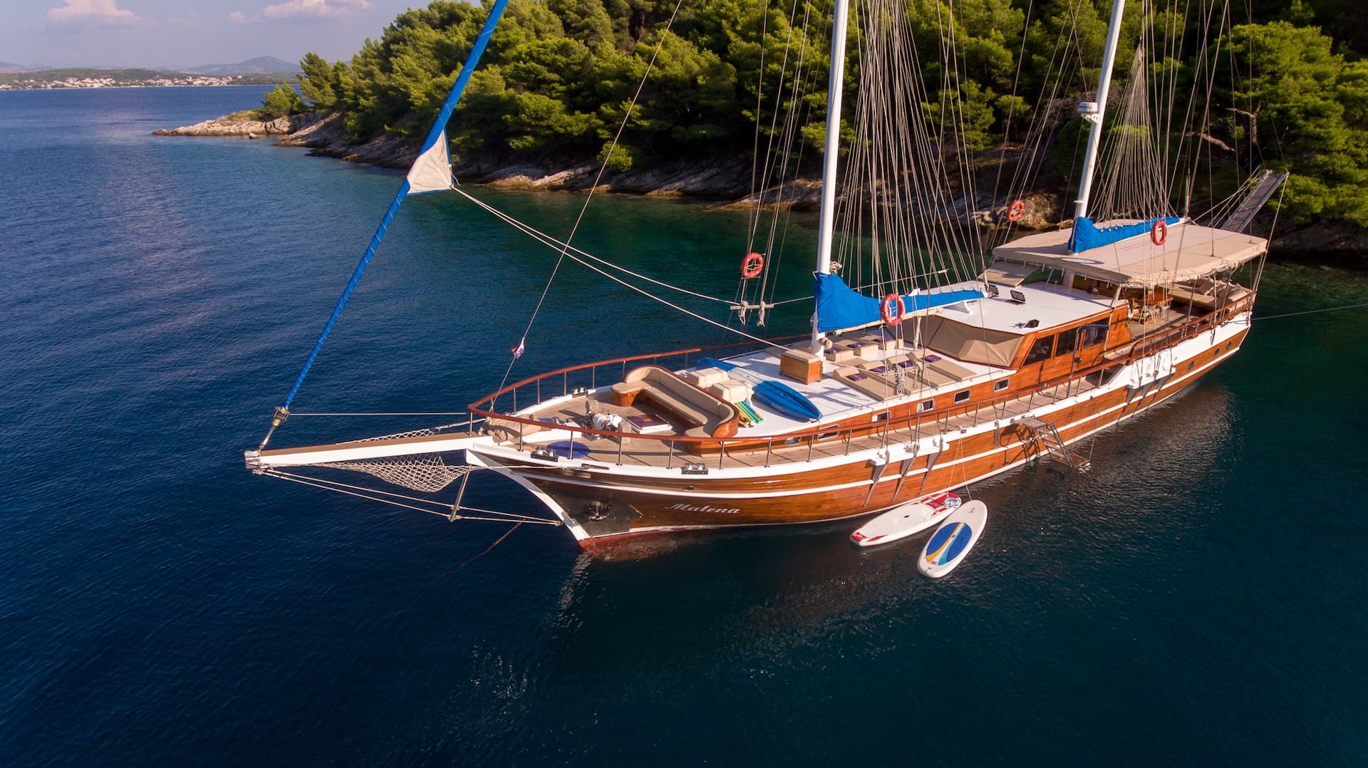 Wooden gulet sailboat named Malena anchored in a cove near a forested coastline.