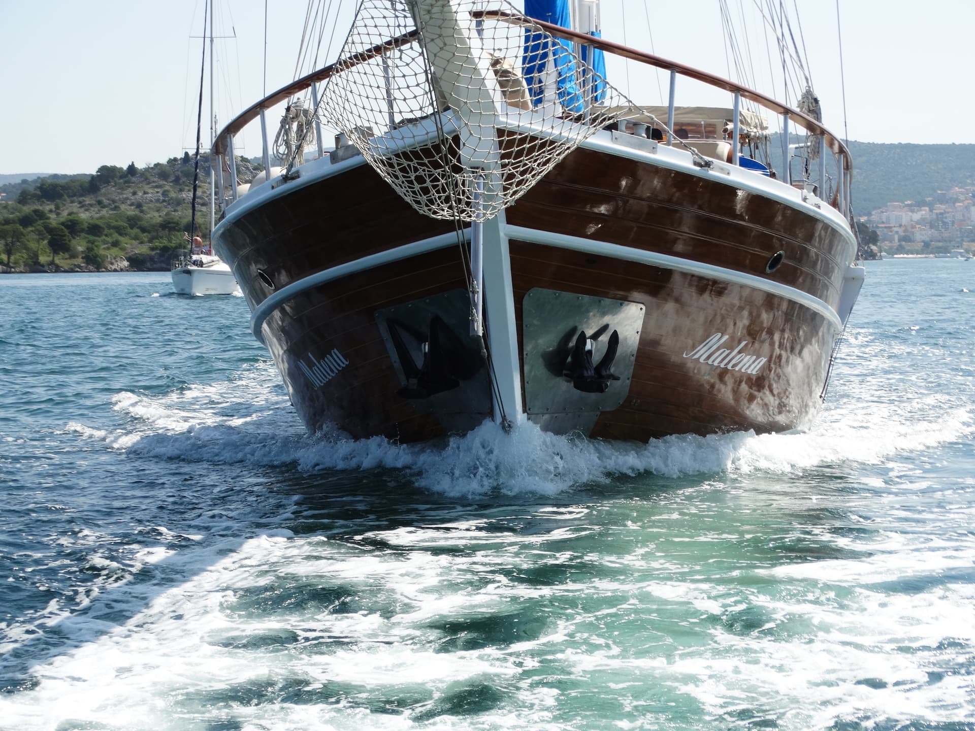 Wooden sailboat named Malena moving through choppy blue water near a coastline.