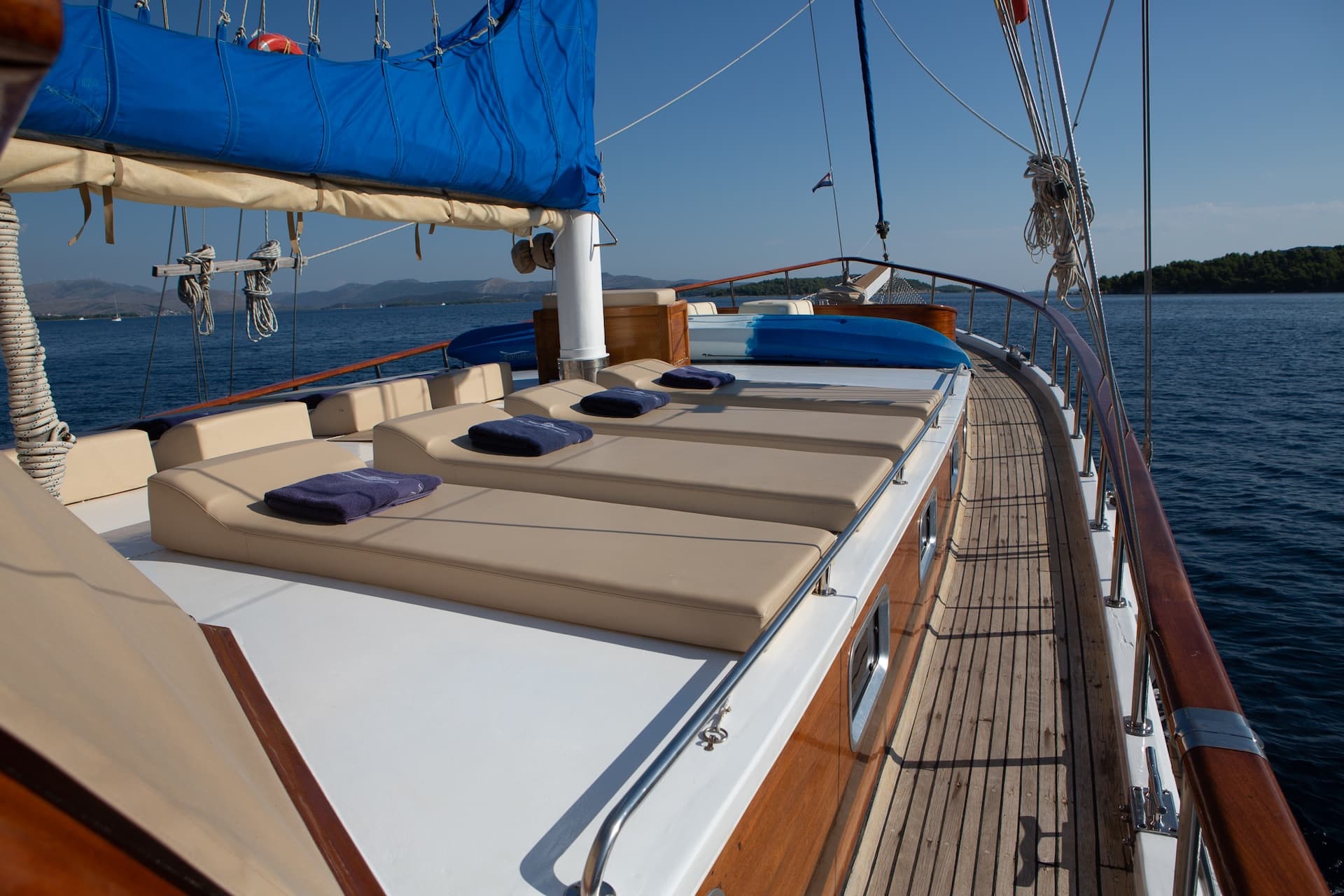 Sun deck with beige loungers and blue towels on a wooden boat sailing near forested islands.