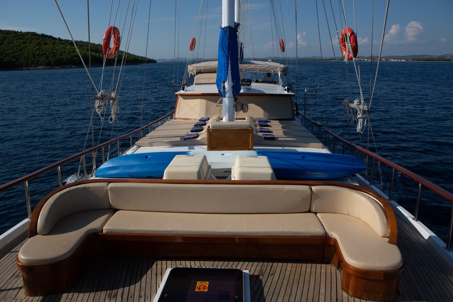 View from sundeck of wooden boat with beige cushions, blue kayaks, and life preservers on blue sea.