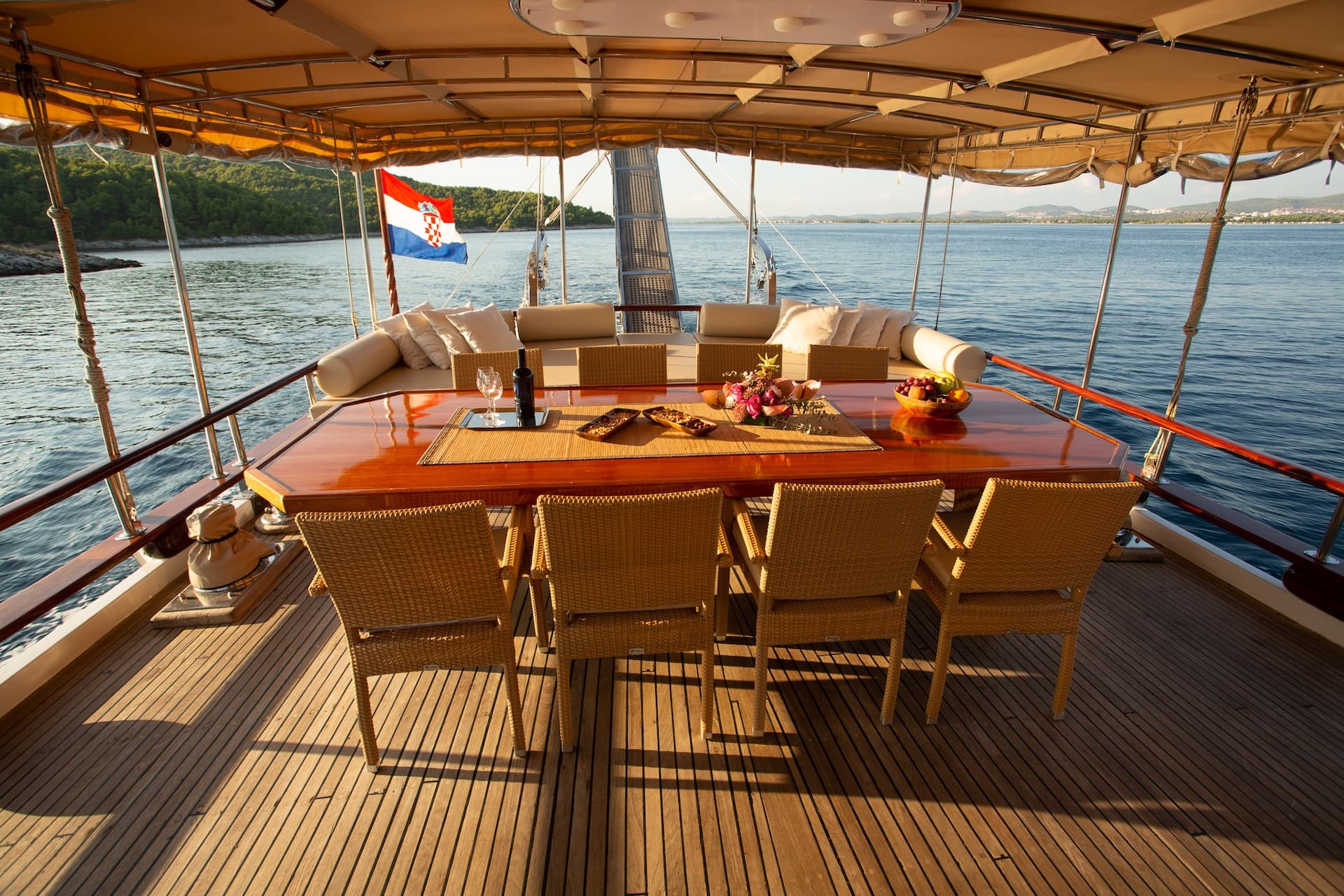 Yacht deck dining area with Croatian flag overlooking wooded coastline and sea.