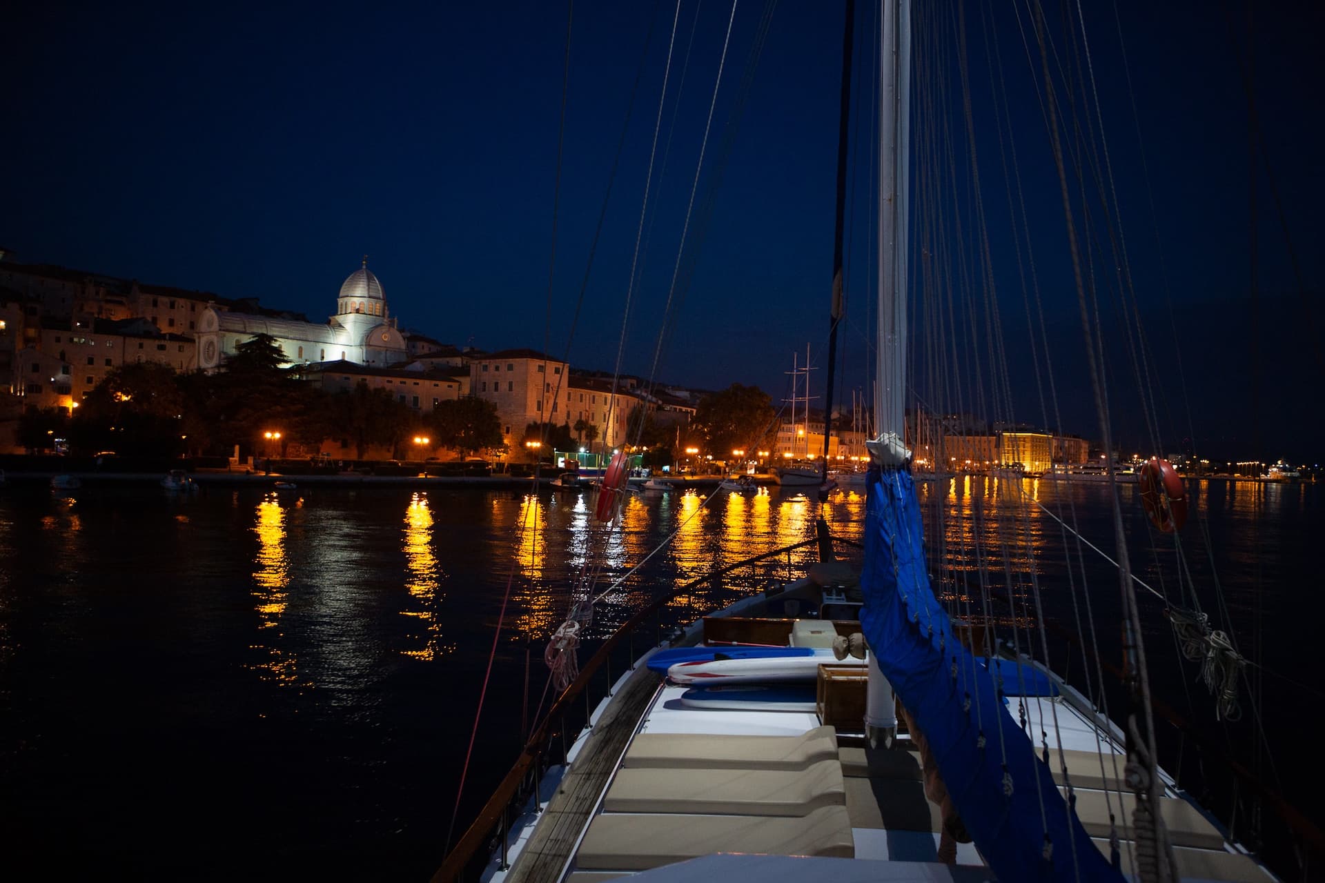 View from sailboat deck at night toward illuminated historic coastal town and harbor.