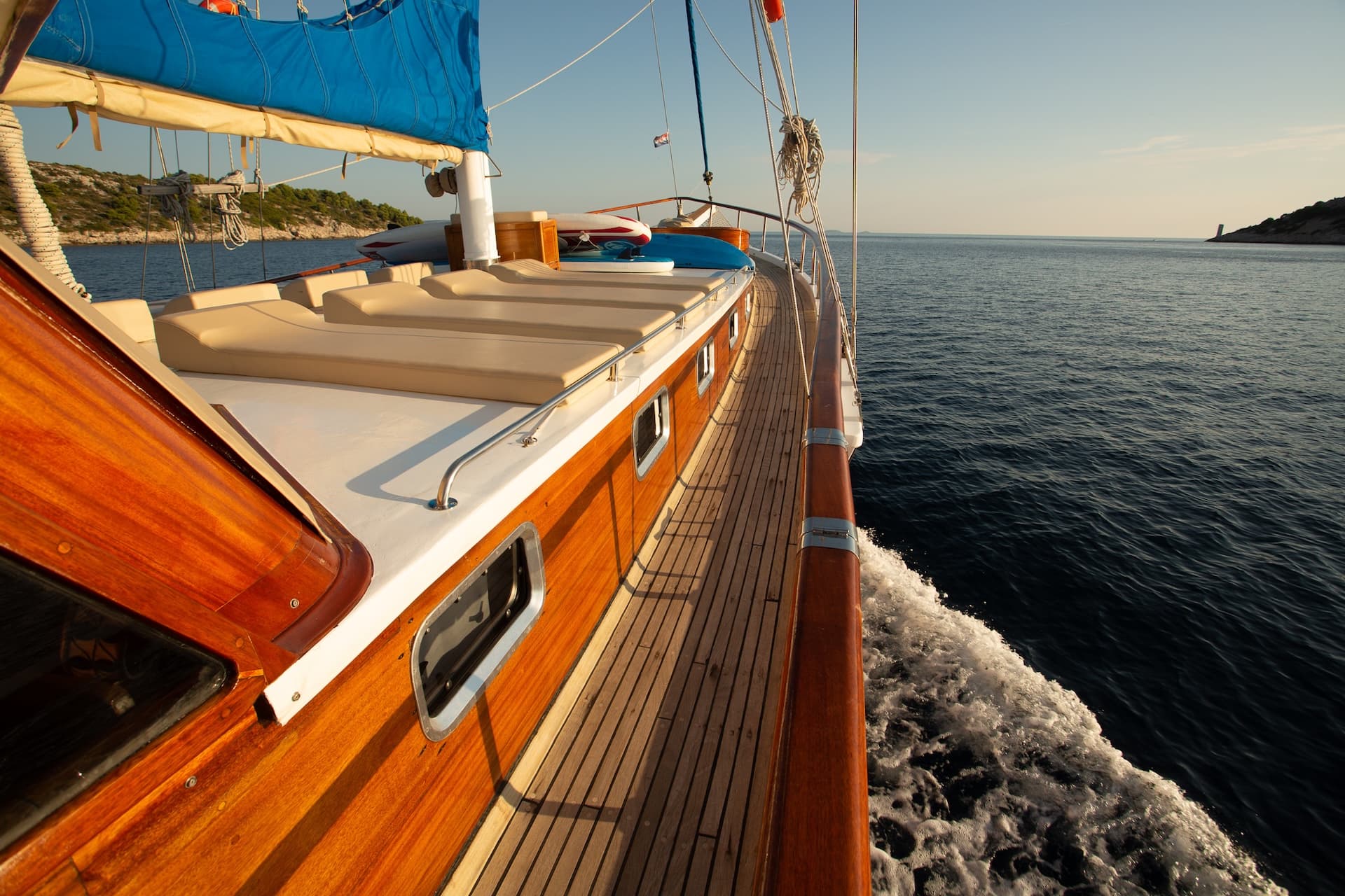 Wooden sailboat moving through dark blue water near a tree-covered coastline.