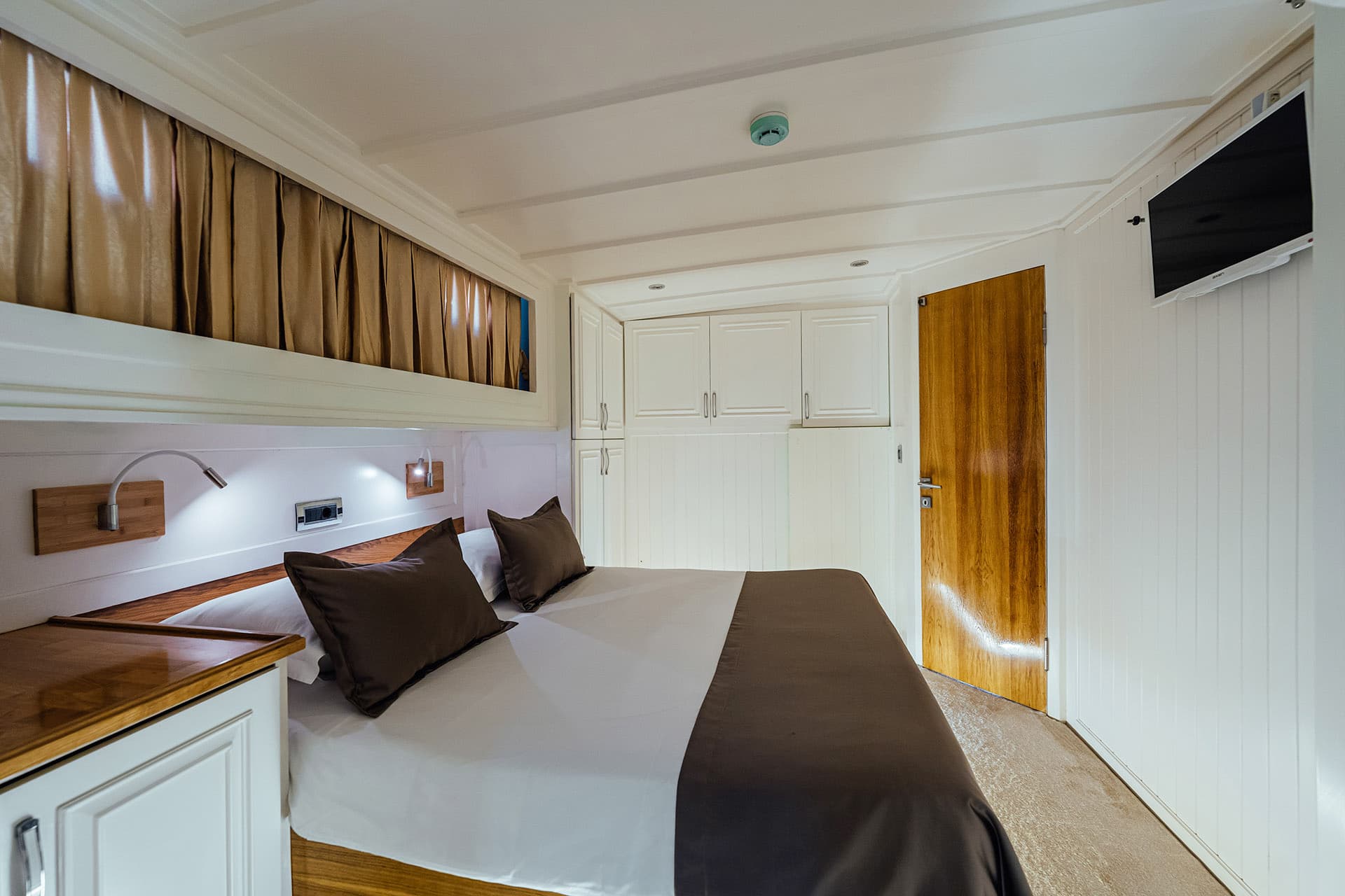 Cosy cabin bedroom with white paneling, wooden door, and brown and white bedding on a Gulet.