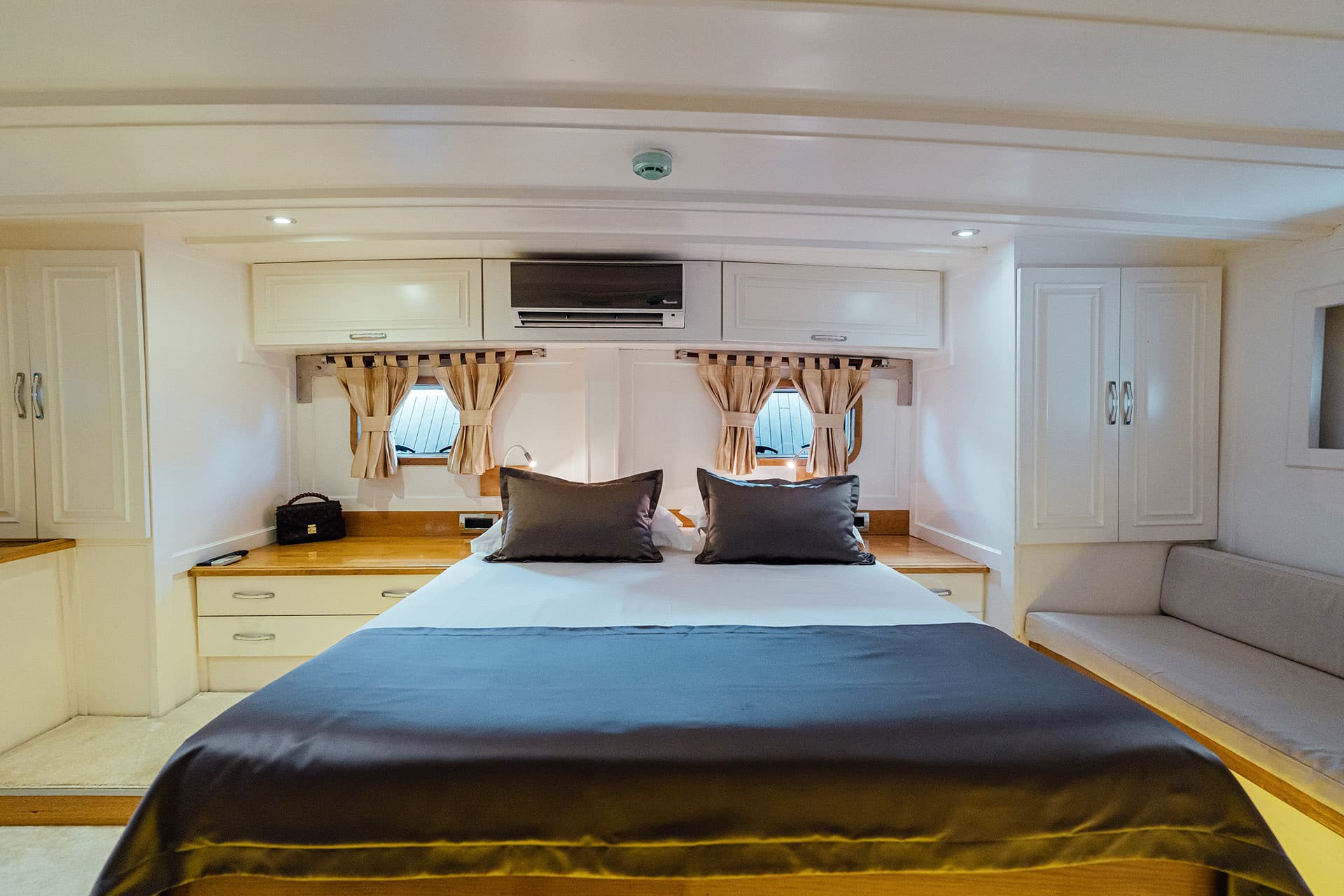 Bedroom cabin interior with double bed, white cabinetry, and porthole windows with curtains.