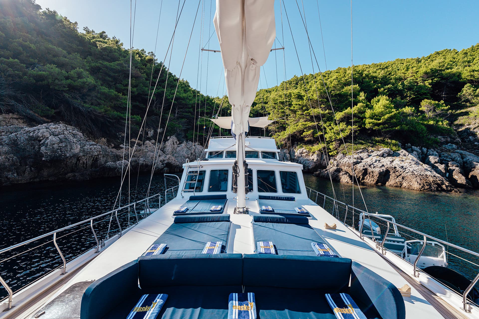 View from bow of yacht with sunbeds anchored near rocky, tree-covered coastline.