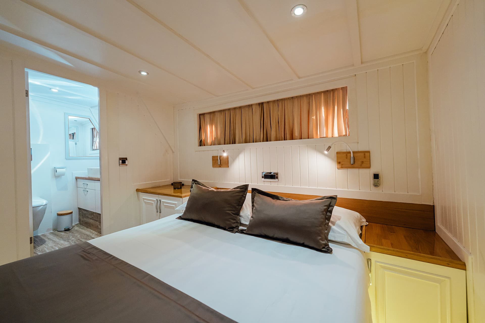 Double cabin interior with white wood paneling, double bed, and view into a bright bathroom.