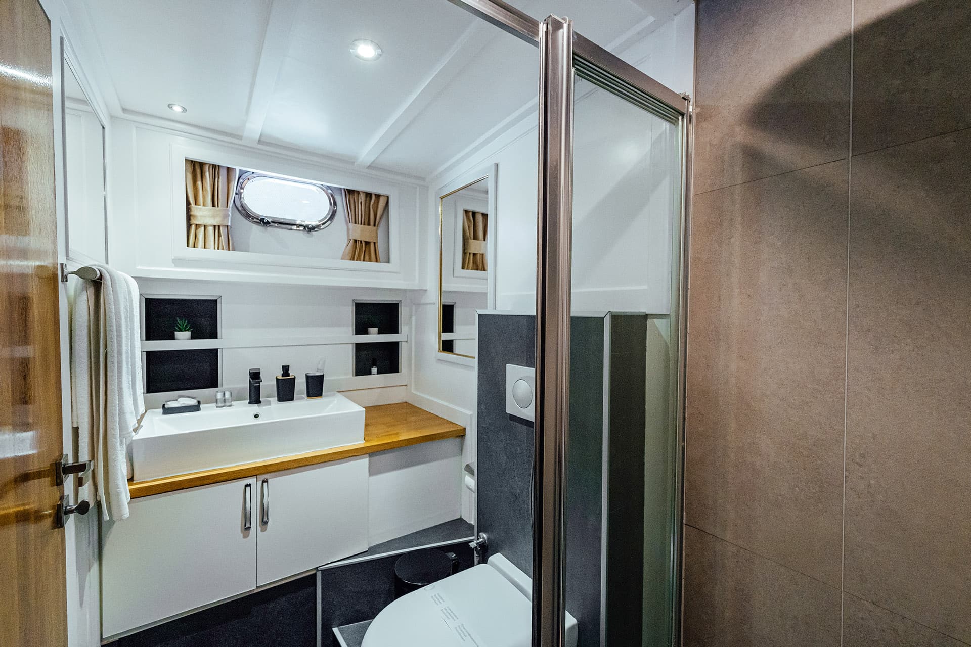 Modern ensuite bathroom on a gulet with white cabinetry, wood counter, and porthole window.
