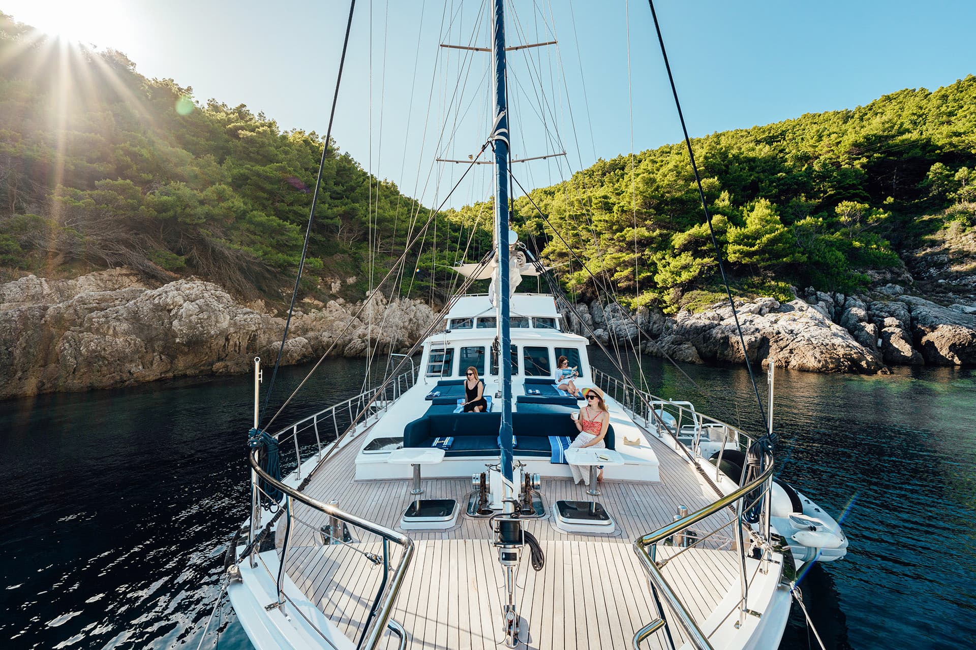 Gulet yacht anchored near rocky, tree-covered coastline with sun flare.