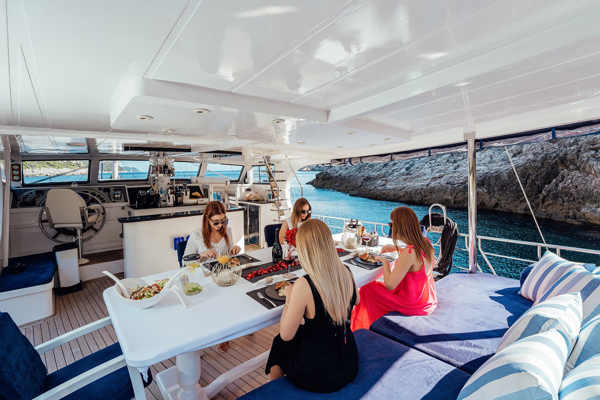 Four guests dining on a yacht deck near rocky coastline with clear blue water