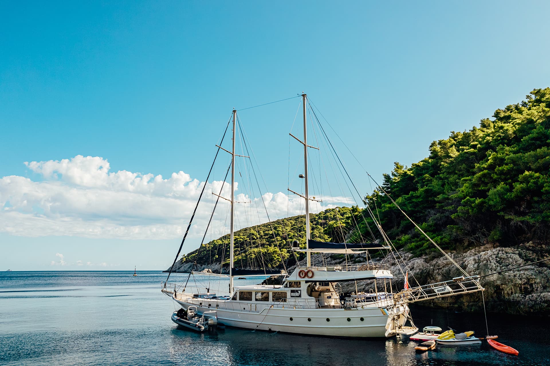 Gulet yacht named Maska moored in calm bay near forested, rocky coastline under blue sky.